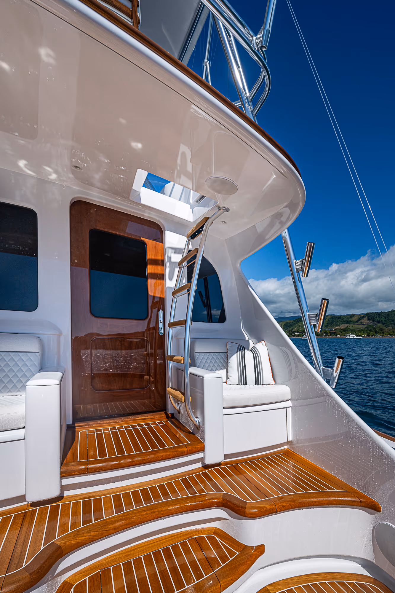 Teak wood steps and seating area with cushions on a white yacht under a clear blue sky near the water.