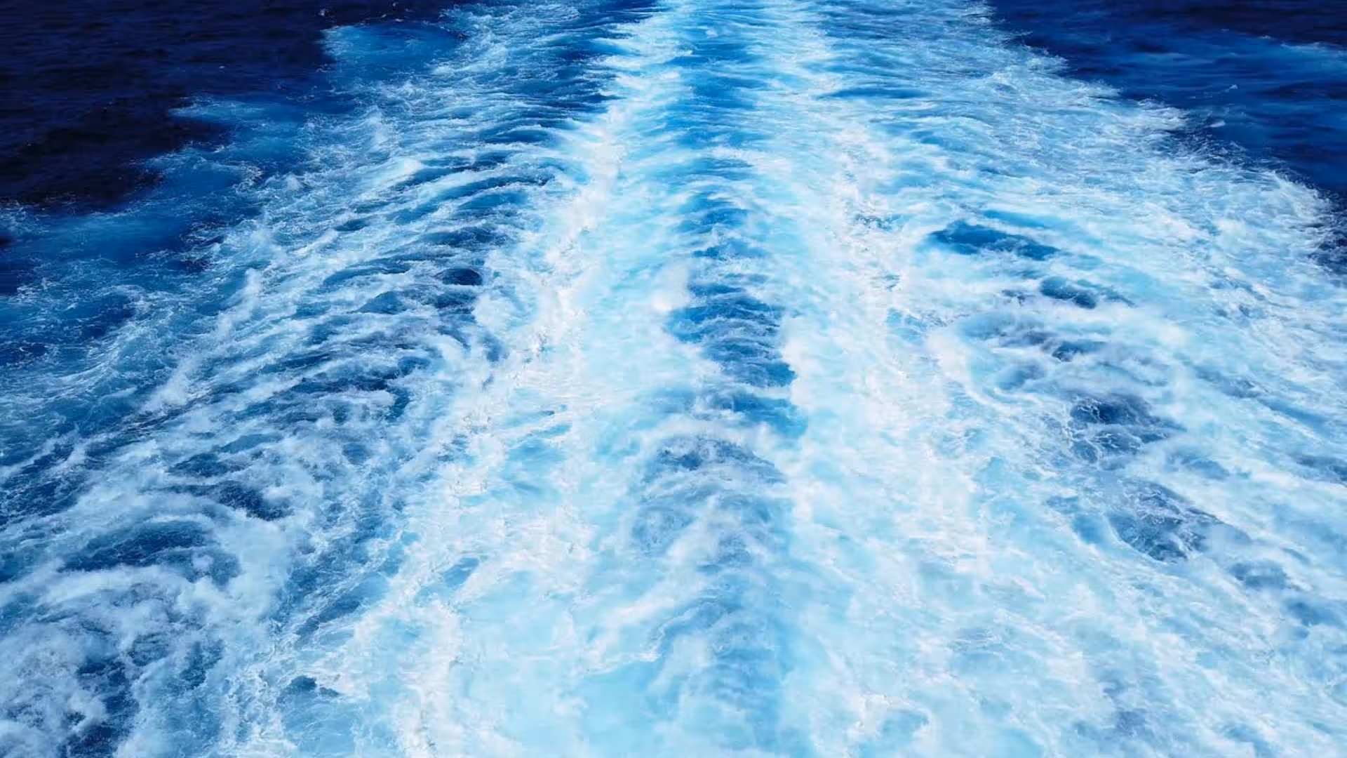 Foamy white and blue water wake trailing behind a moving boat on deep blue ocean.
