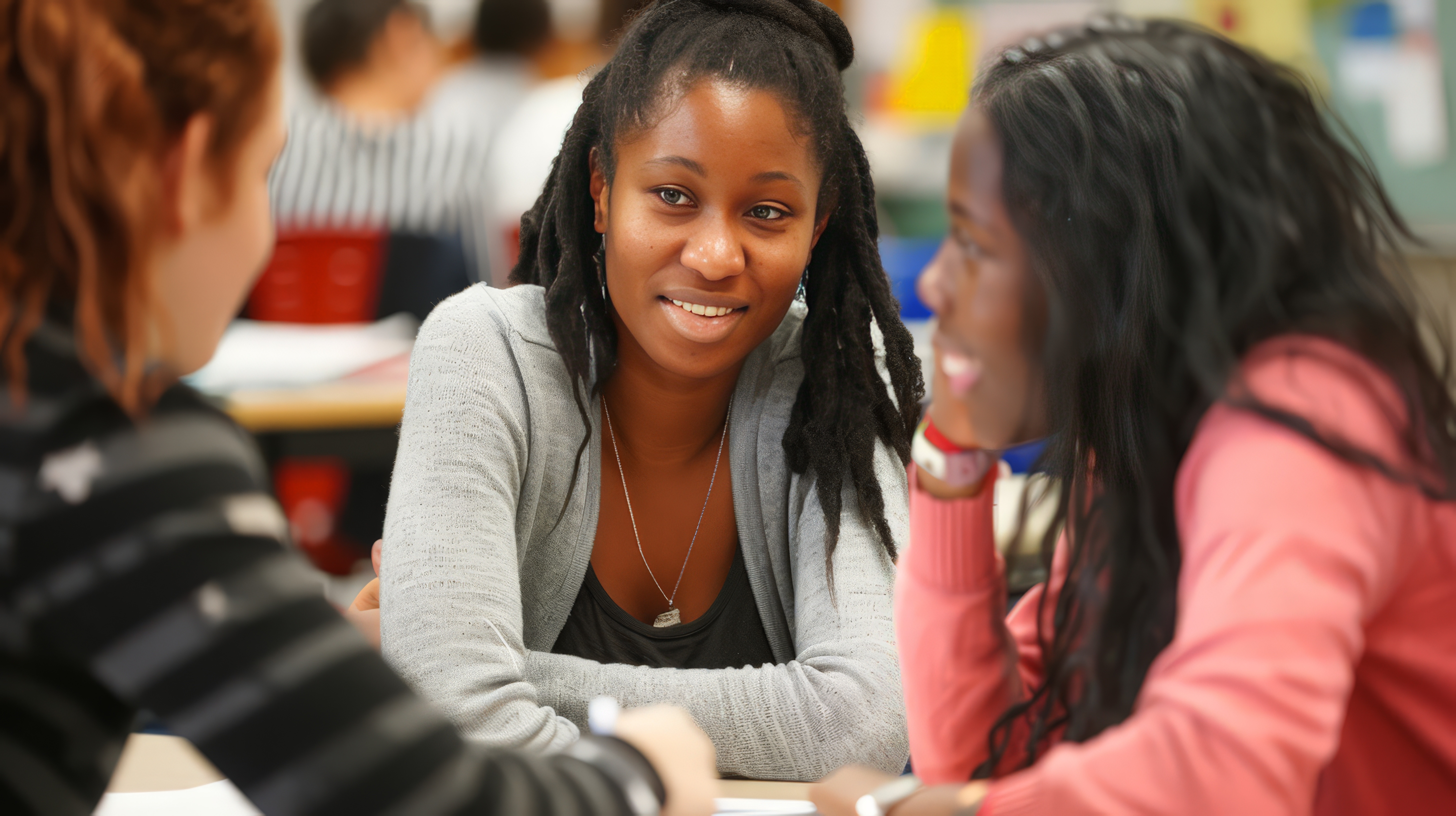 Three women engaged in a conversation at a table in a classroom setting.