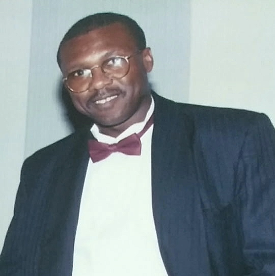 Man wearing glasses, a dark suit, white shirt, and a red bow tie smiling in front of a plain background.
