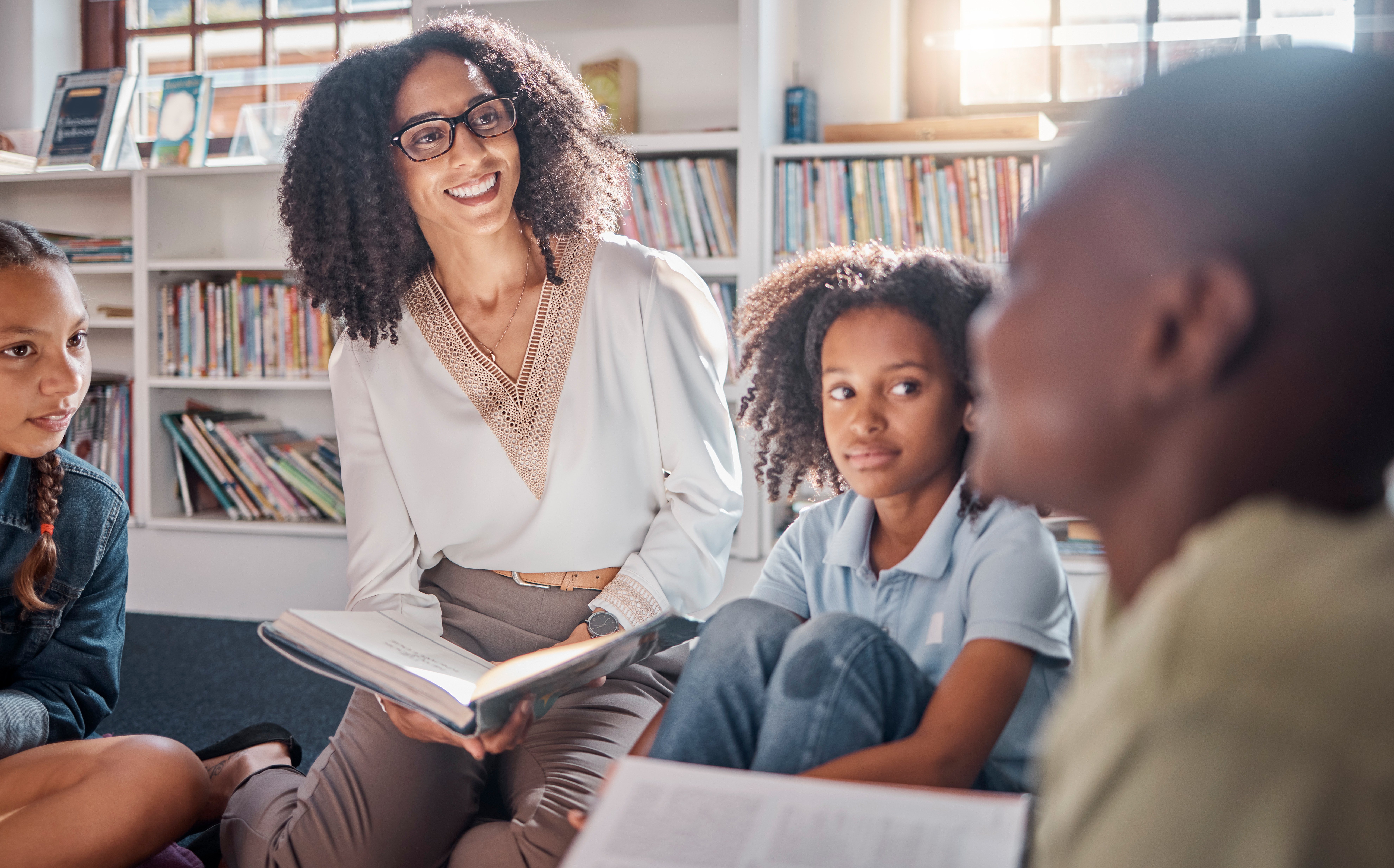 Teacher with glasses smiling and reading a book to children sitting on the floor in a bright library.