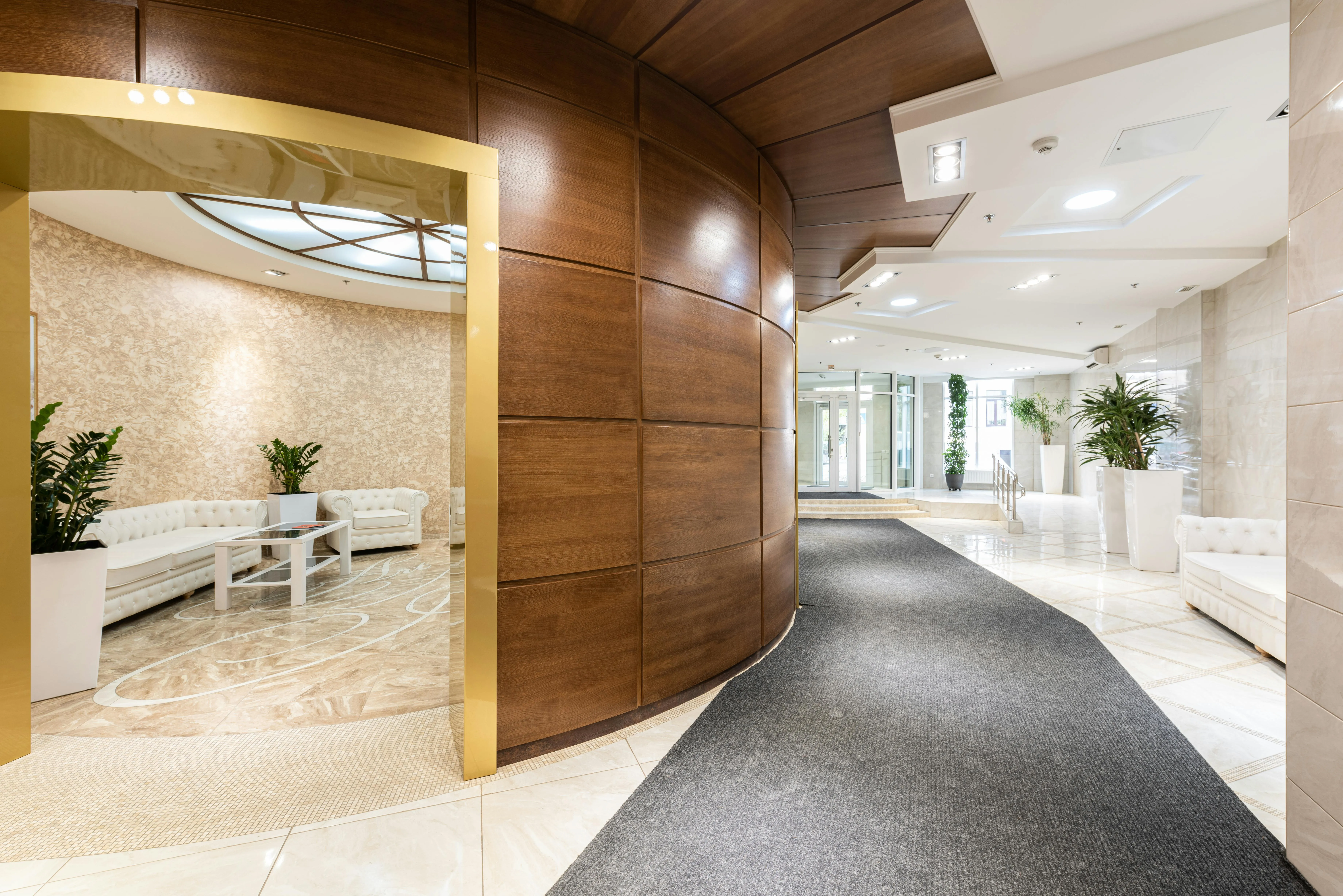 A wide shot of a modern, bright hotel lobby and reception area. A curved wall with polished wood panelling separates a cozy seating area on the left—which features white tufted sofas, a coffee table, and green plants—from a long hallway with light marble flooring and a grey carpet runner leading toward glass entrance doors.