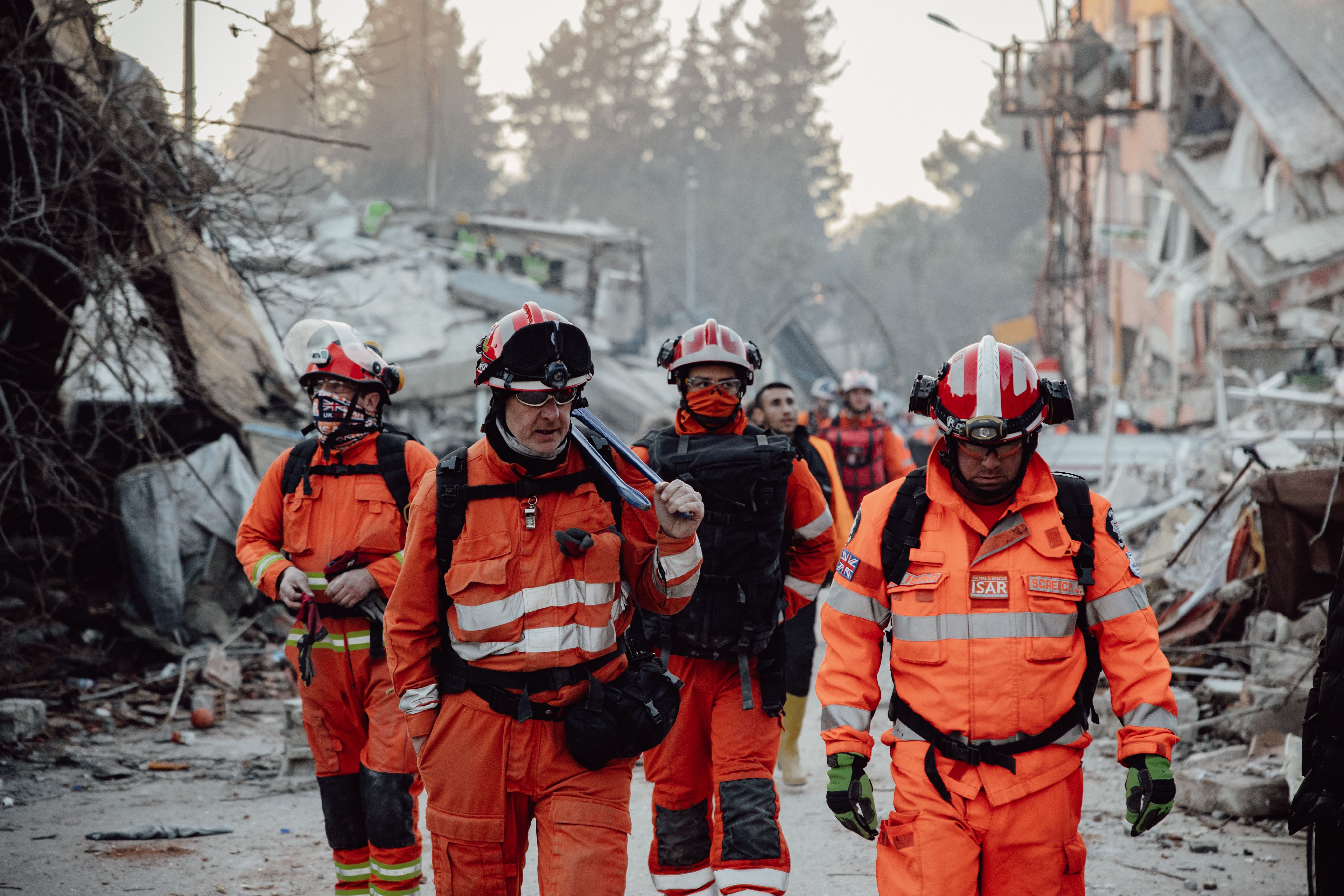 Search and rescue workers wearing orange protective suits and helmets walking through rubble at a disaster scene.