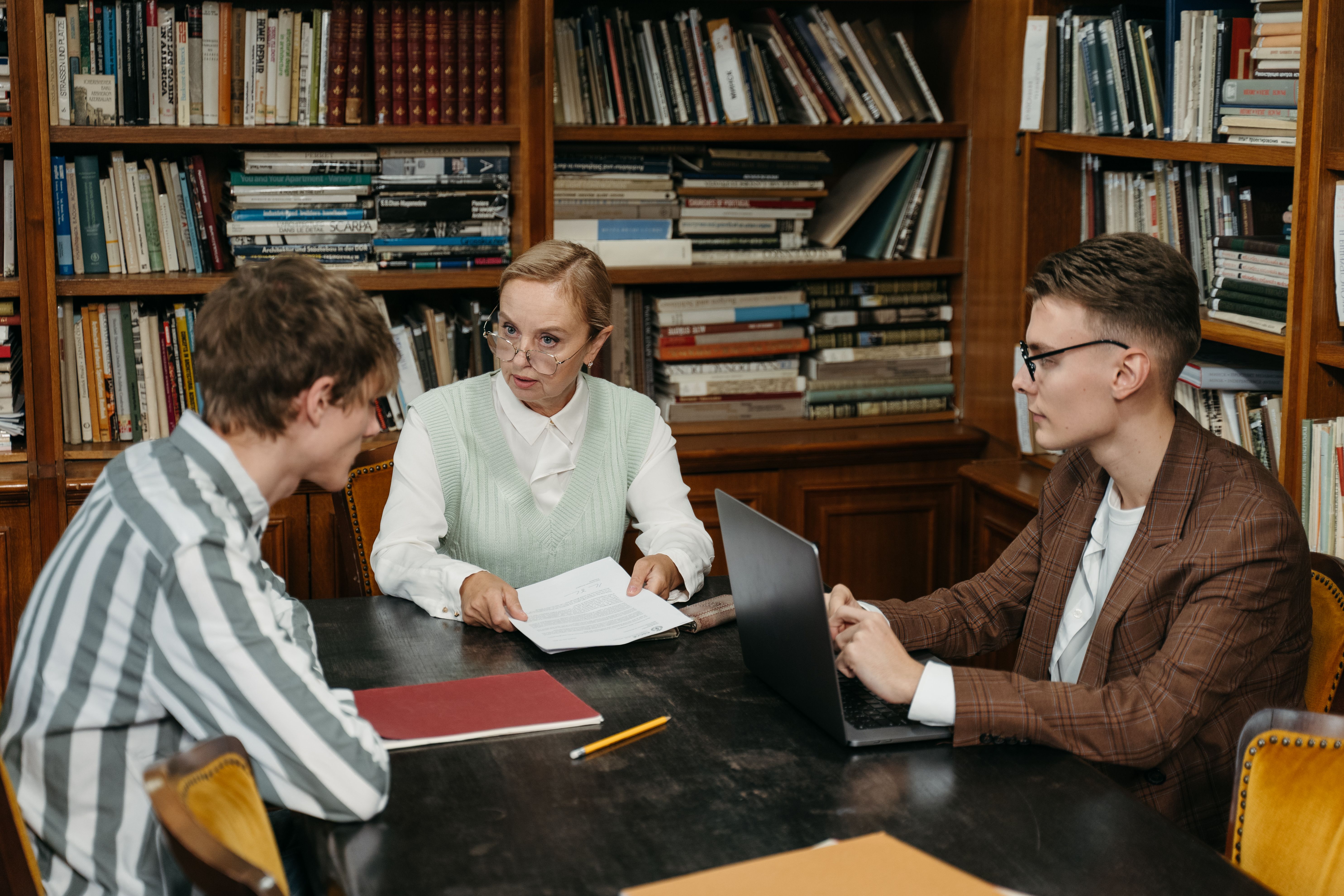 An instructor in glasses holding documents while discussing coursework with two male students seated at a wooden desk in a library.