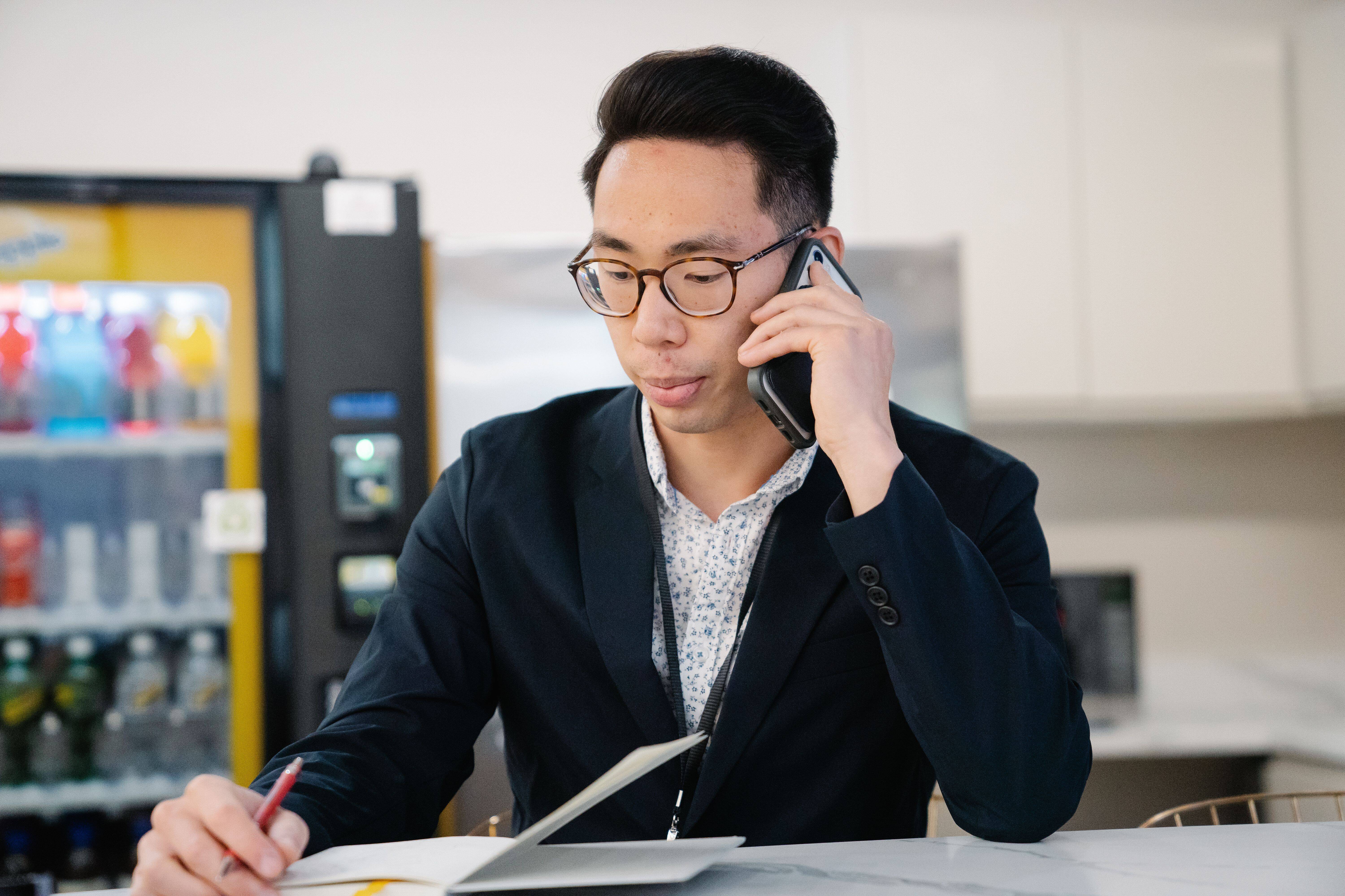 A man in a suit jacket and glasses writing in a notebook while talking on his mobile phone near a breakroom vending machine.