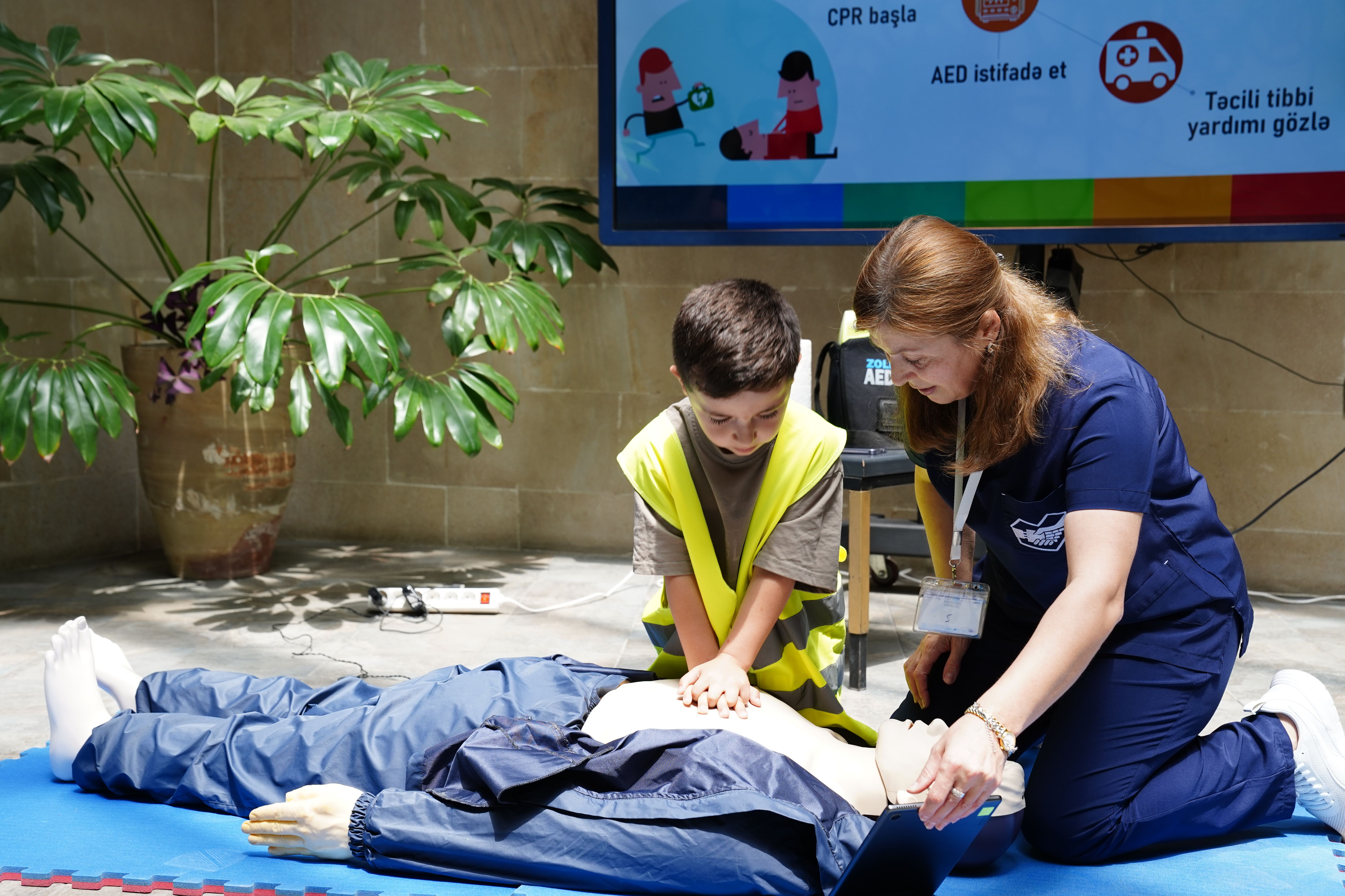 An instructor in scrubs guiding a young boy in a high-visibility vest through hands-on CPR training on a CPR mannequin.