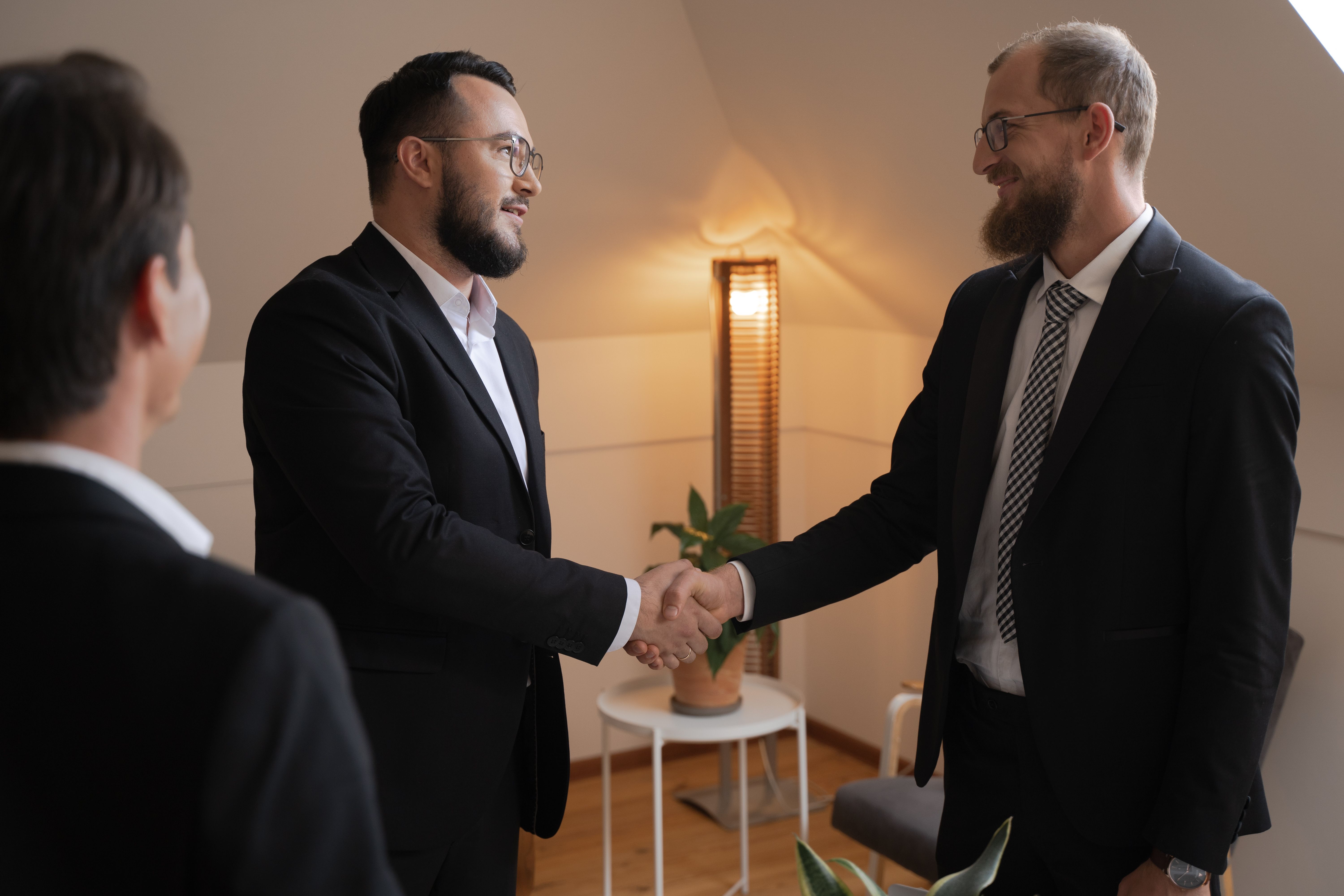 Two businessmen in suits smiling and shaking hands during a professional corporate meeting indoors.
