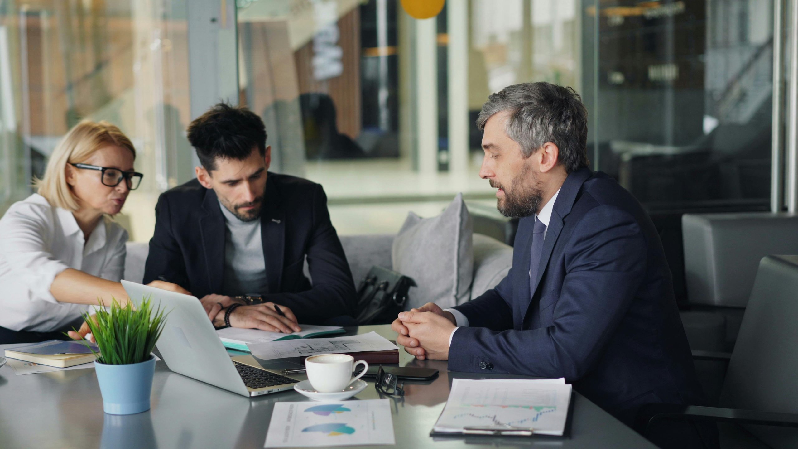 Three business professionals reviewing documents and charts at a conference table with a laptop and coffee cup.