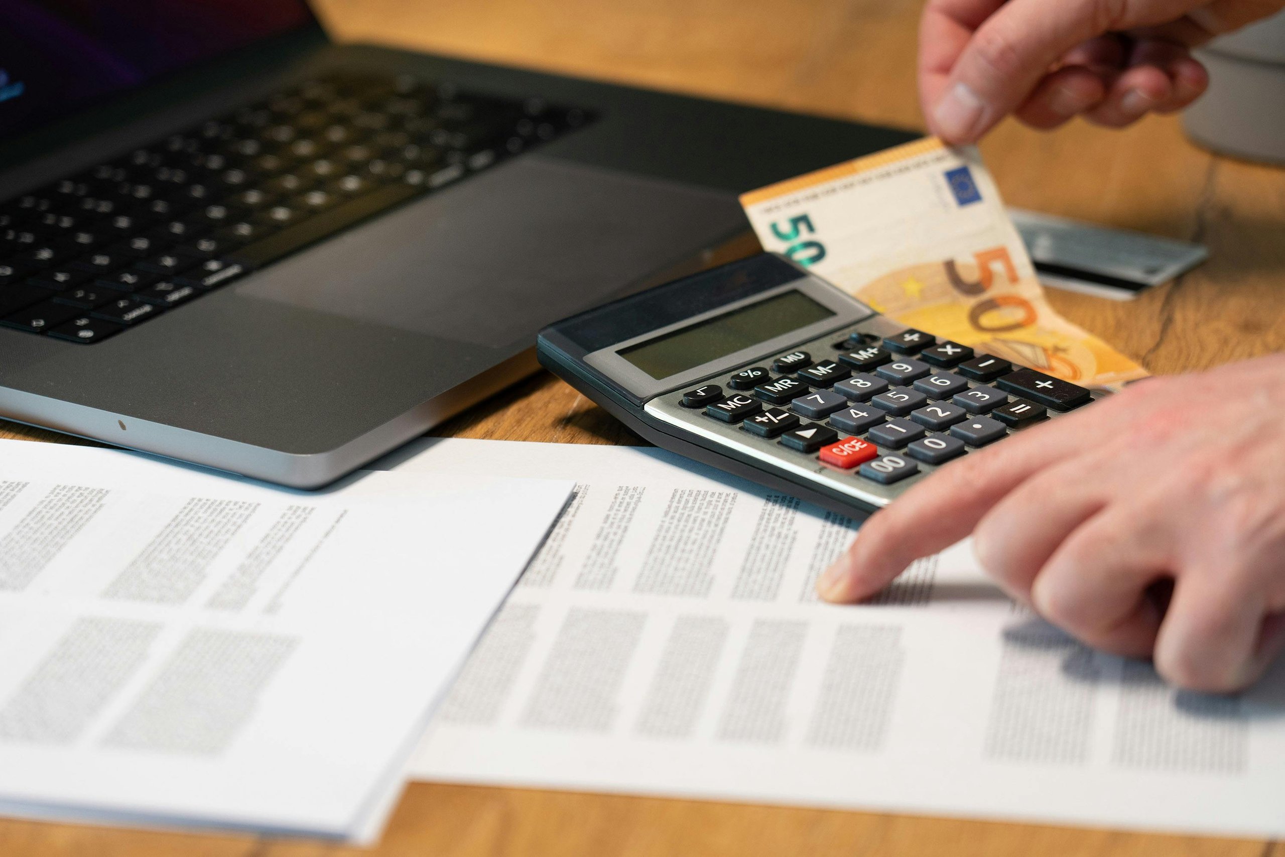 Person using calculator and pointing at financial documents next to a laptop and holding a 50 euro banknote.