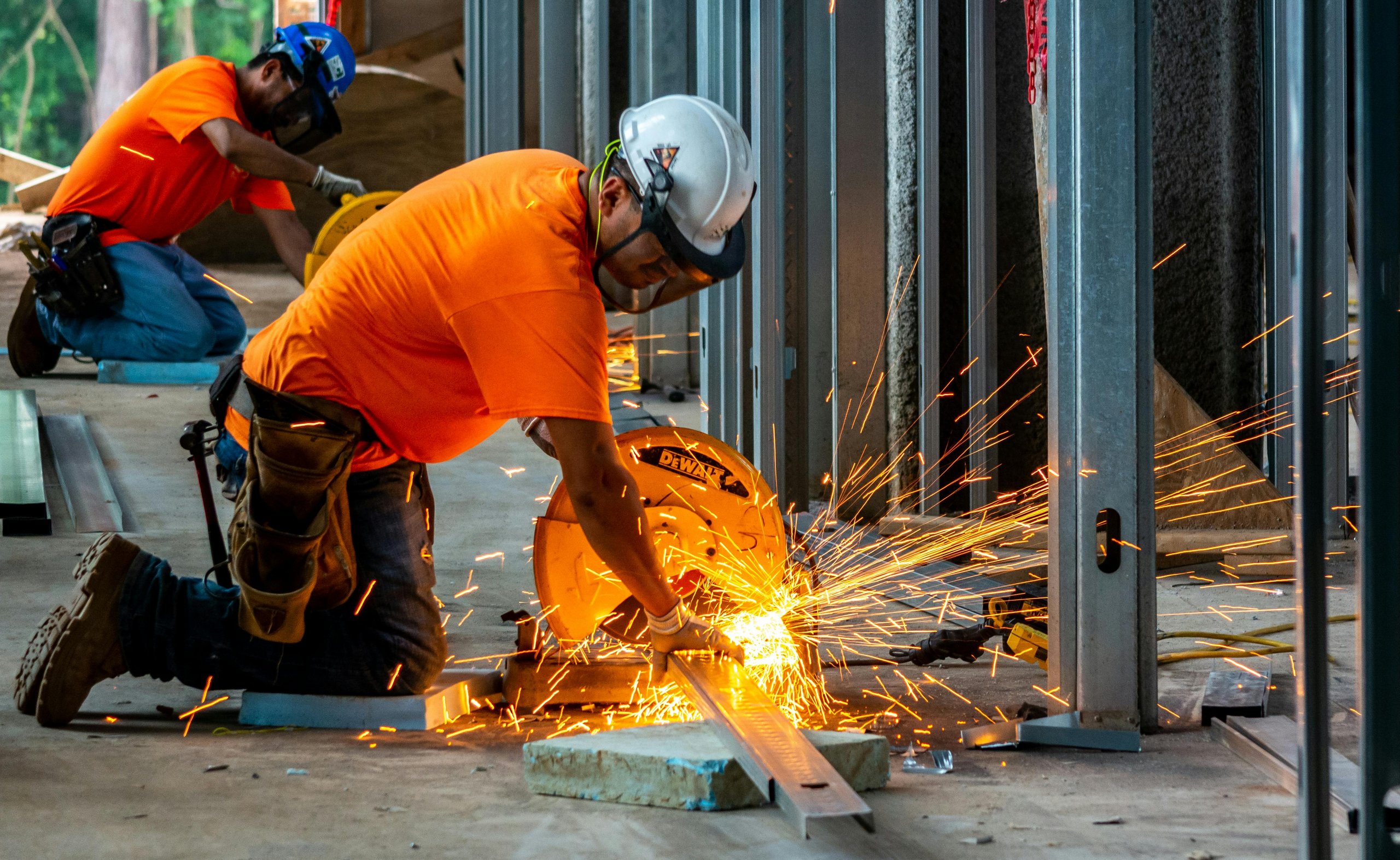 Two construction workers in orange shirts and helmets cutting metal with a circular saw, sparks flying.