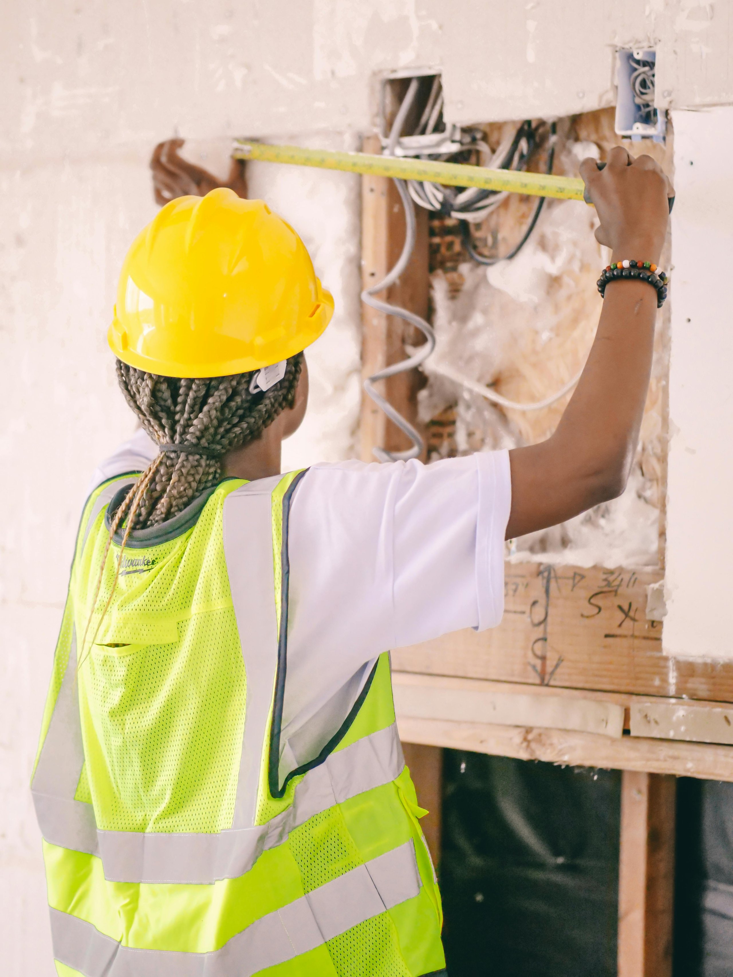Construction worker wearing a yellow hard hat and reflective vest measures an open wall section with a tape measure.