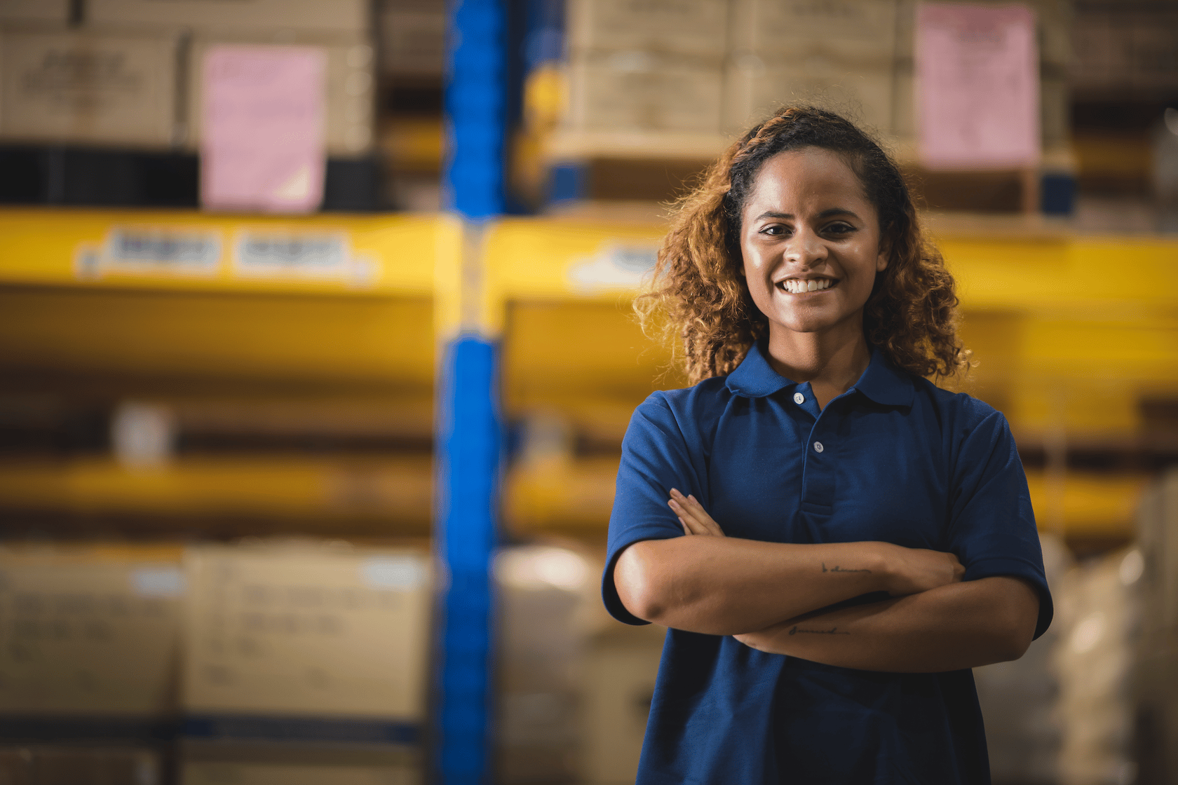 Smiling woman standing with crossed arms in a warehouse filled with stacked boxes and yellow shelving.