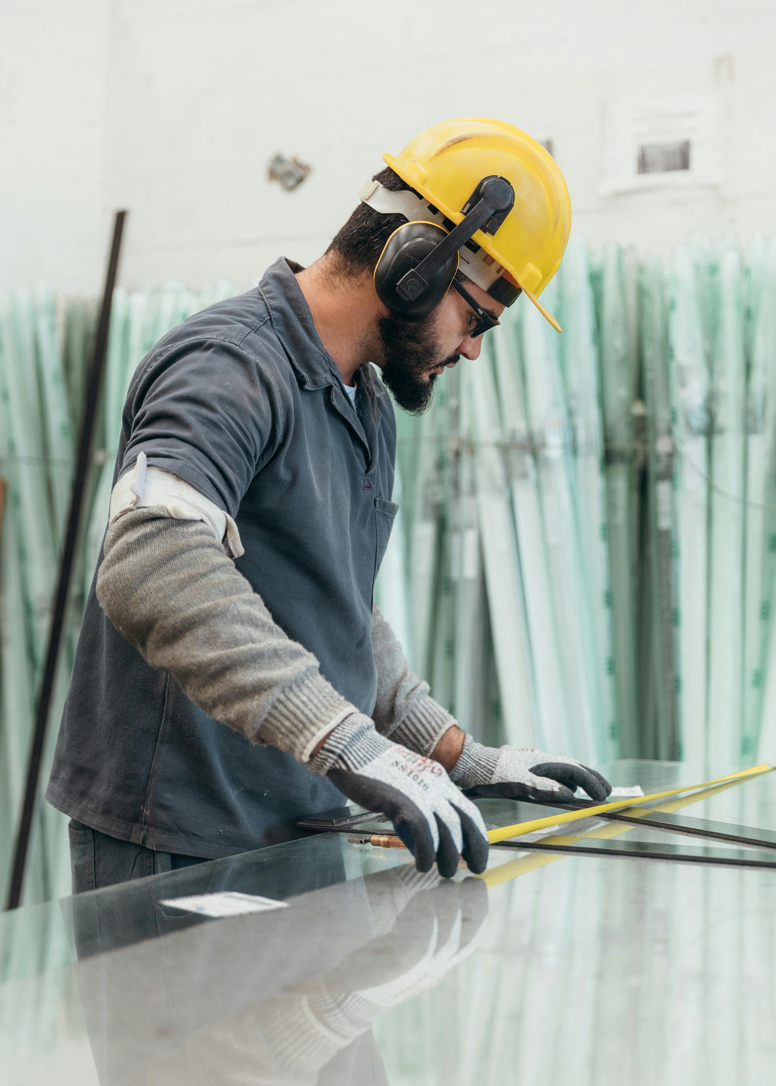 Construction worker wearing a yellow hard hat and gloves measuring glass with a tape measure.