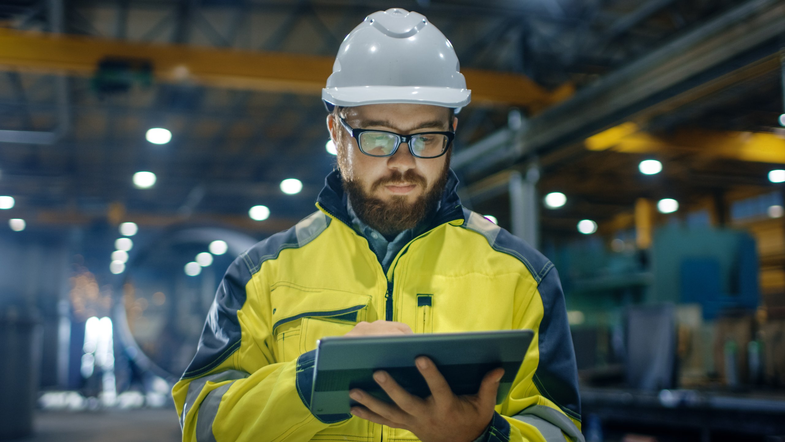 Worker in a yellow safety jacket and white hard hat using a tablet inside an industrial facility.