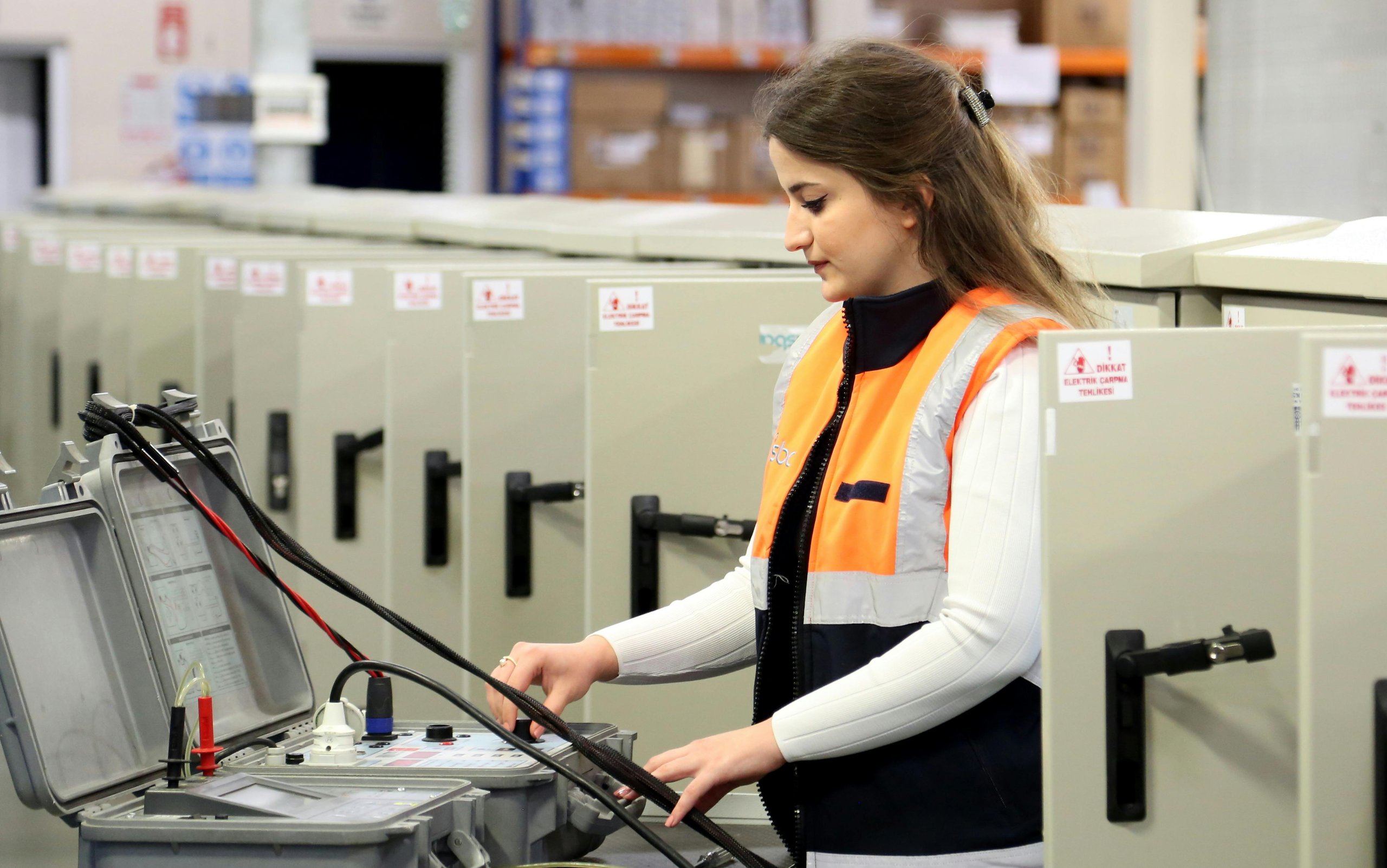 Woman in an orange safety vest working with electrical testing equipment in an industrial setting.
