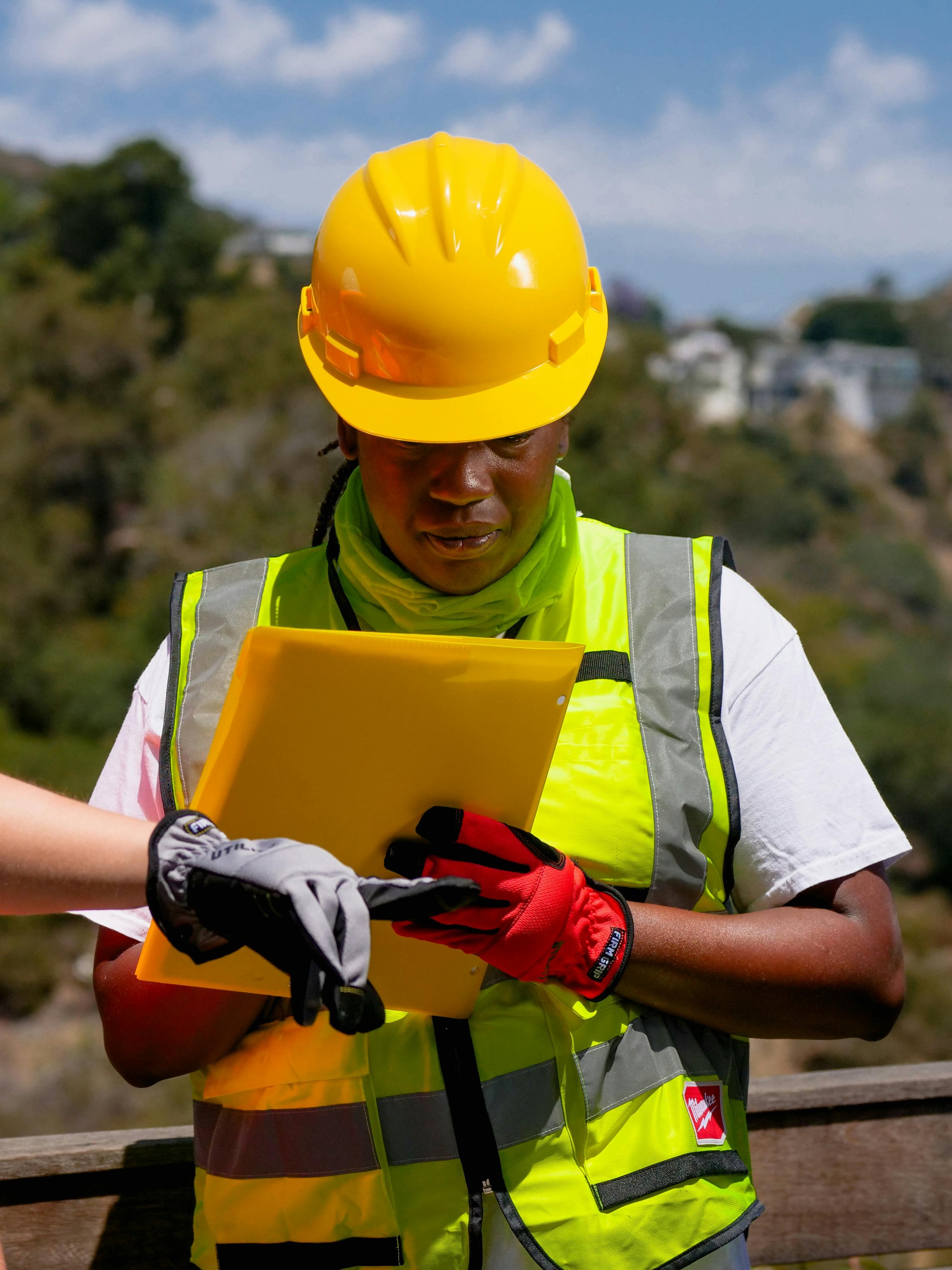 Construction worker wearing a yellow hard hat and safety vest reviewing documents on a yellow clipboard while another gloved hand points.