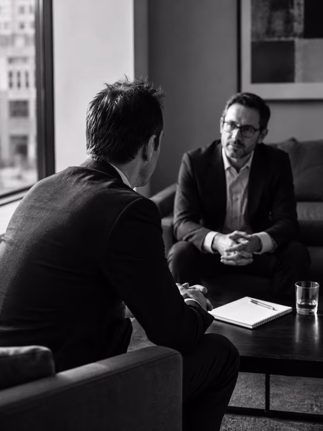 Two men in suits having a serious conversation in a modern office with notebook and glass on table.