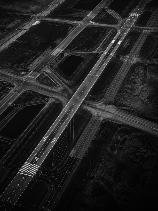 Black and white aerial view of an empty airport runway and intersecting taxiways with markings.
