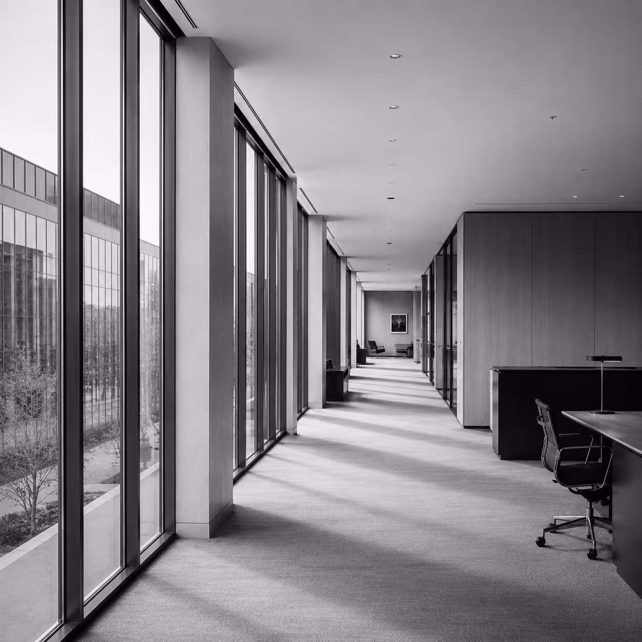 Sunlit modern office corridor with floor-to-ceiling windows and minimalist furniture in grayscale.