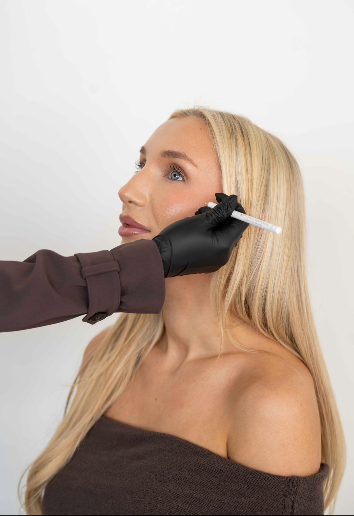 Person with black-gloved hand holding a white cosmetic pencil near a blonde woman's cheek for makeup application.