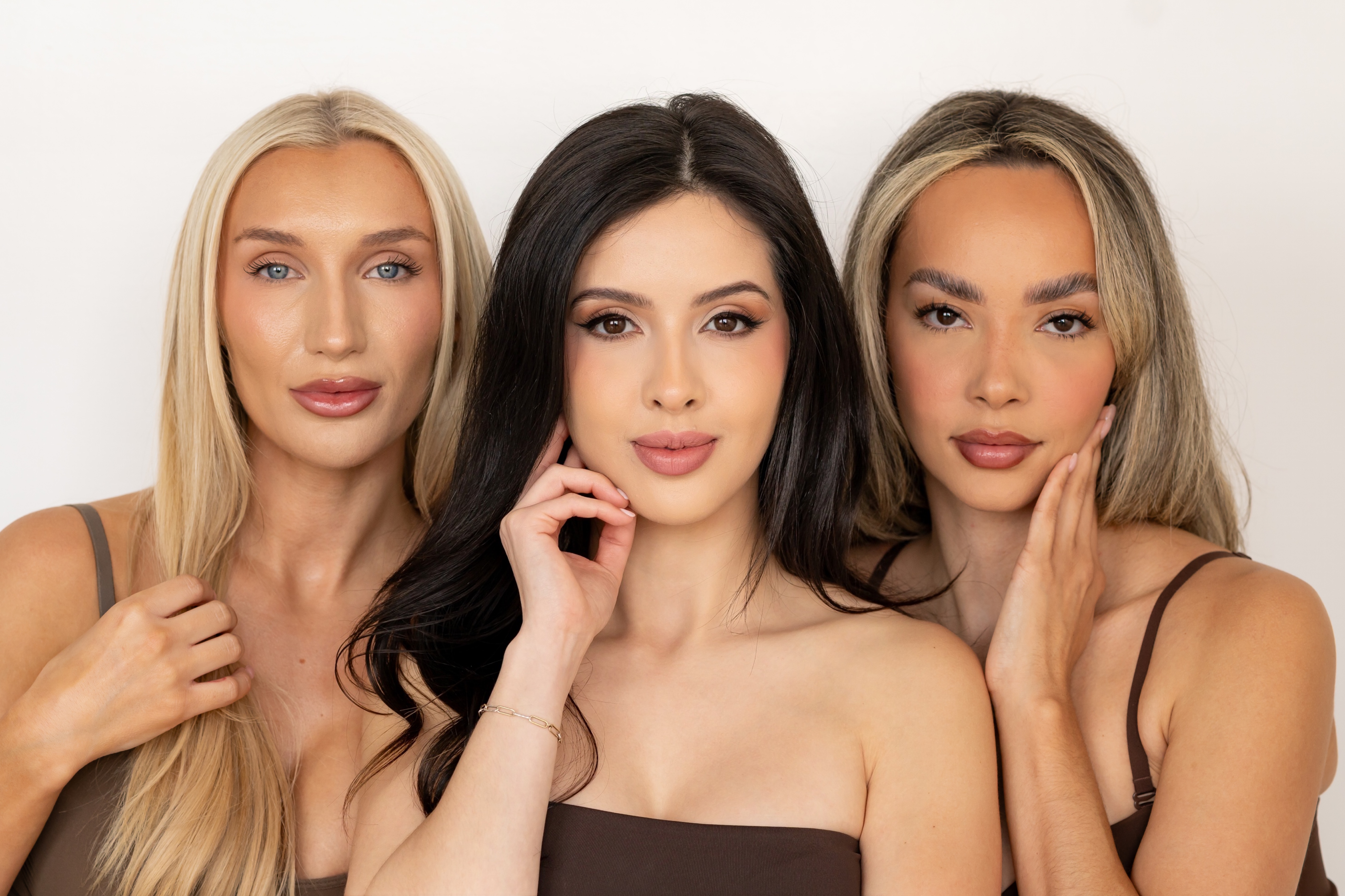 Portrait of three women with different hair colors and natural makeup wearing brown tops against a white background.