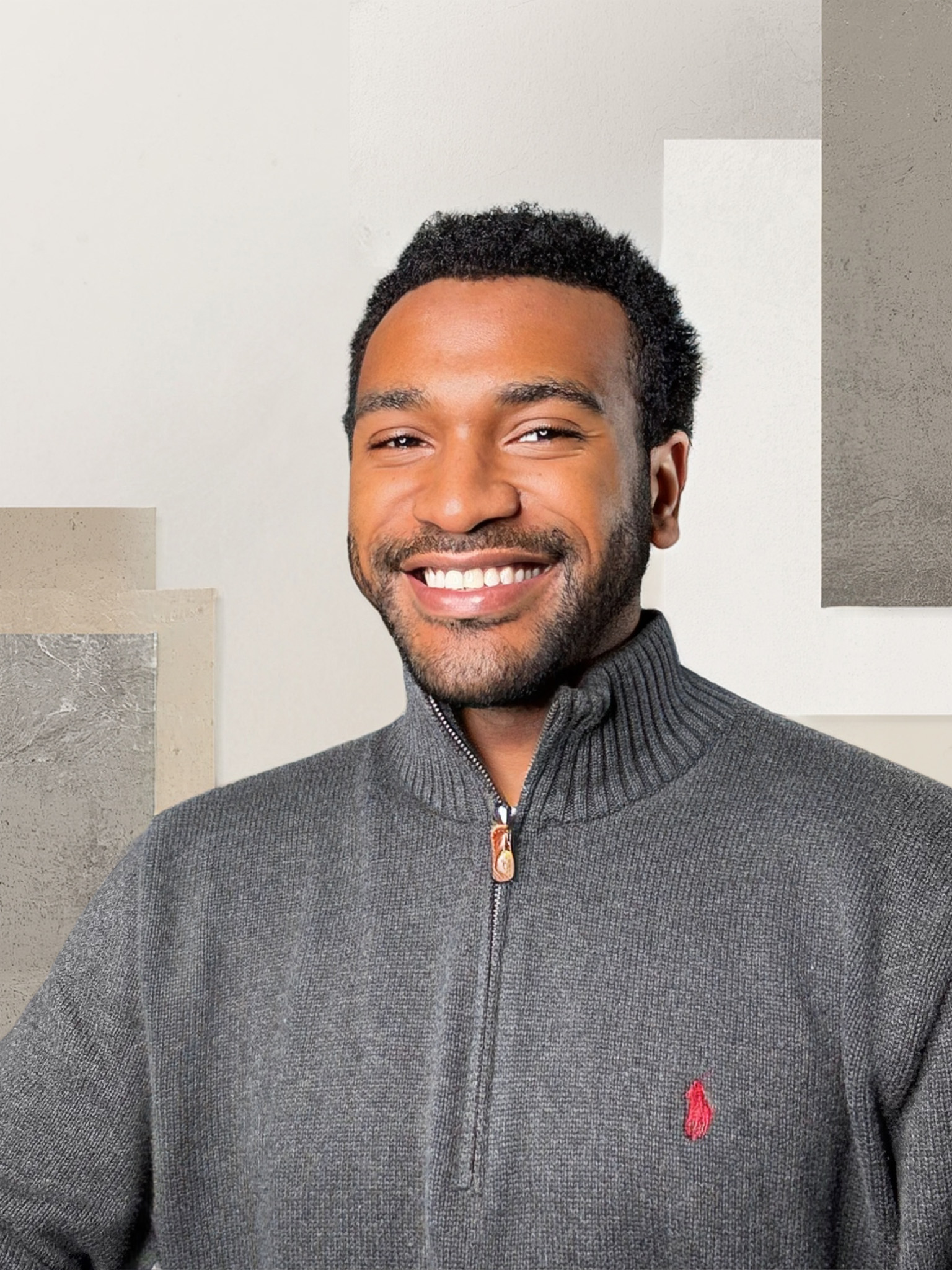 Smiling man with short curly hair wearing a gray zip-up sweater with a red embroidered logo.