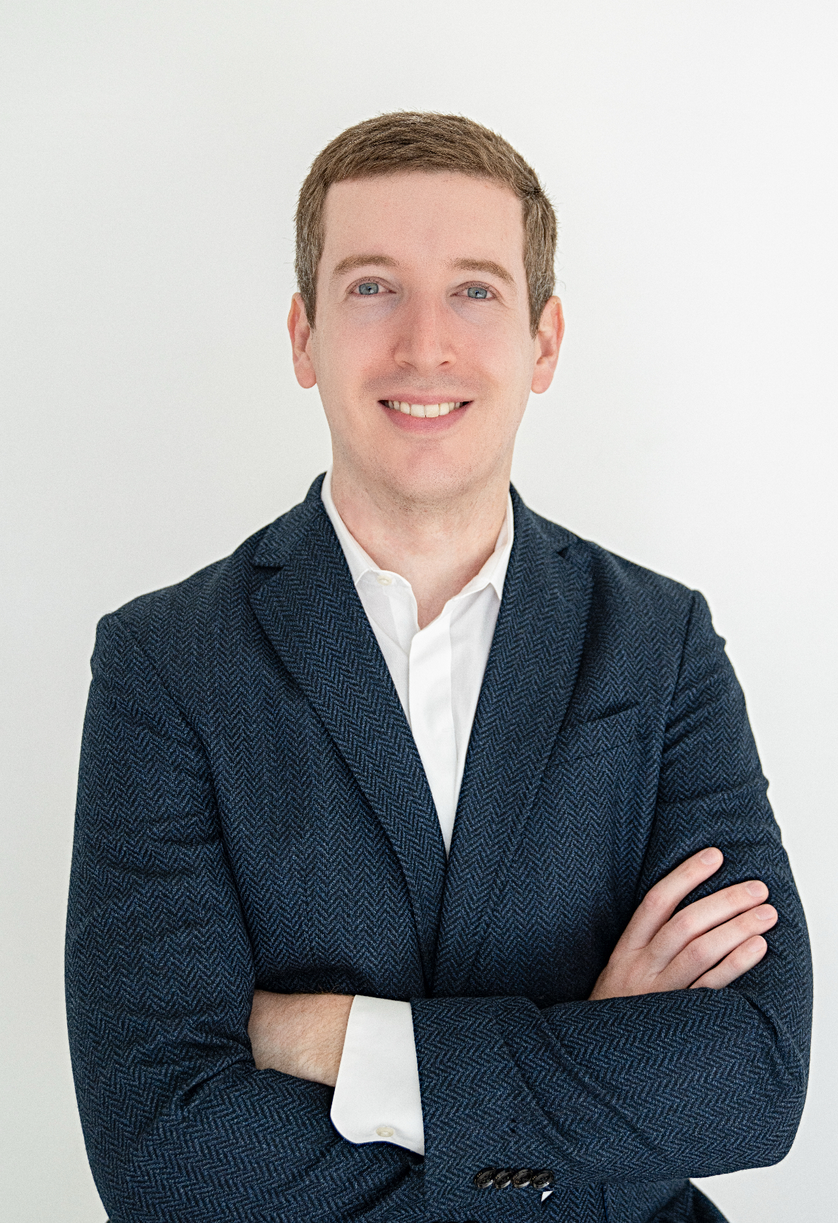 Smiling man with short brown hair wearing a white shirt and dark blue blazer, arms crossed against a plain white background.