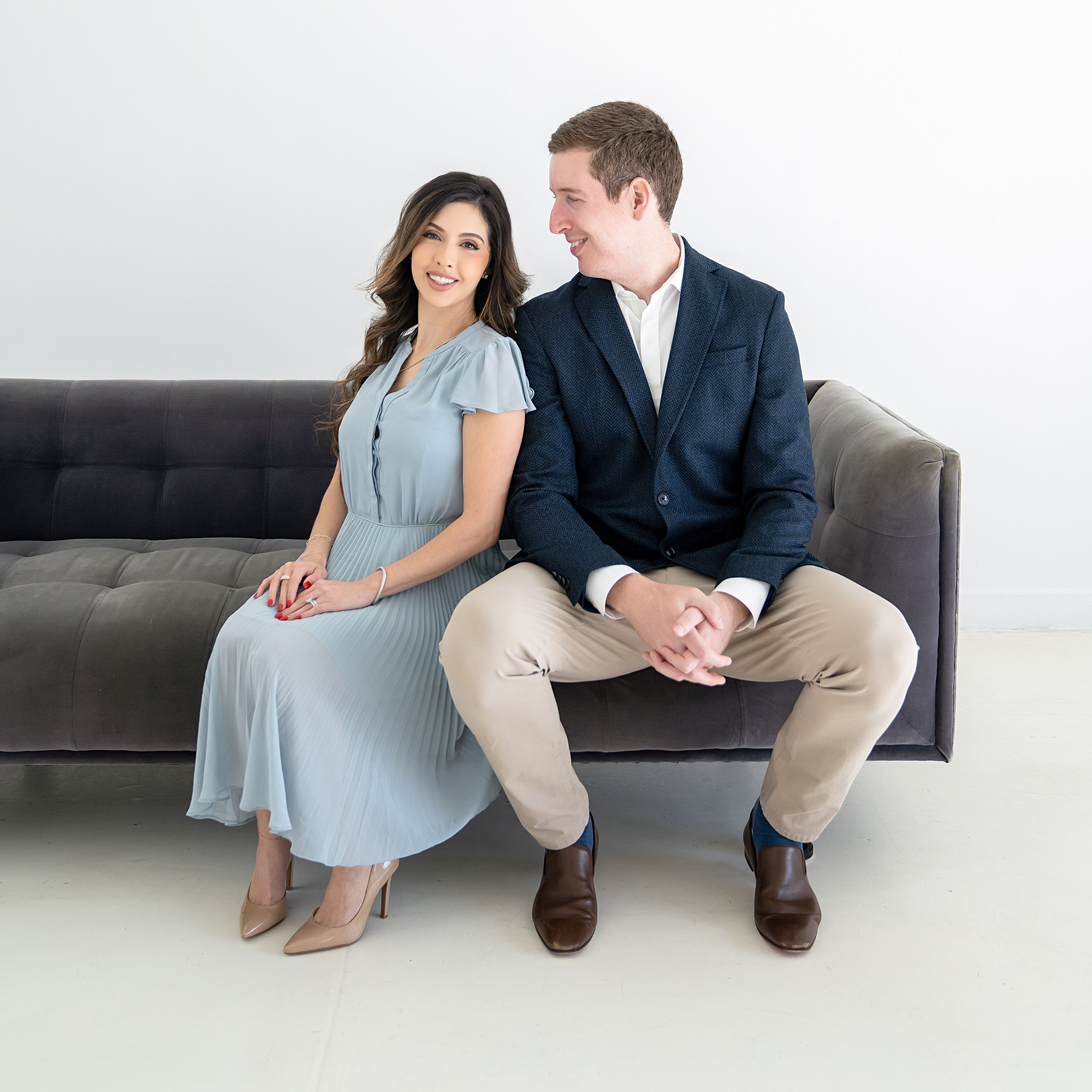 Woman in a light blue dress and man in a dark blazer sitting on a gray sofa against a white background.