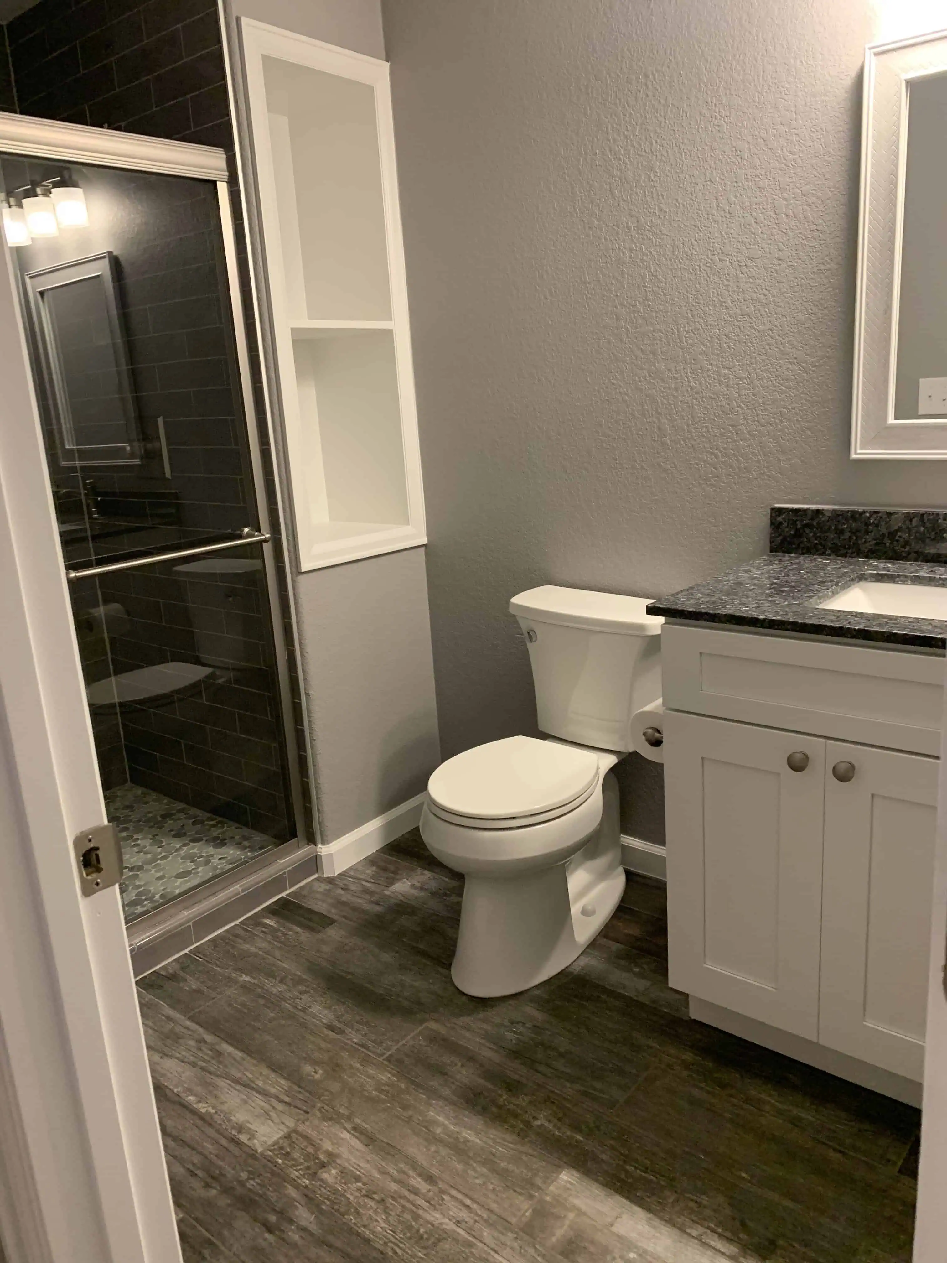 Modern bathroom renovation by Blue Mountain Remodeling in Denver, featuring a sleek shower with glass doors, a granite countertop, white cabinetry, and wood-look tile flooring.
