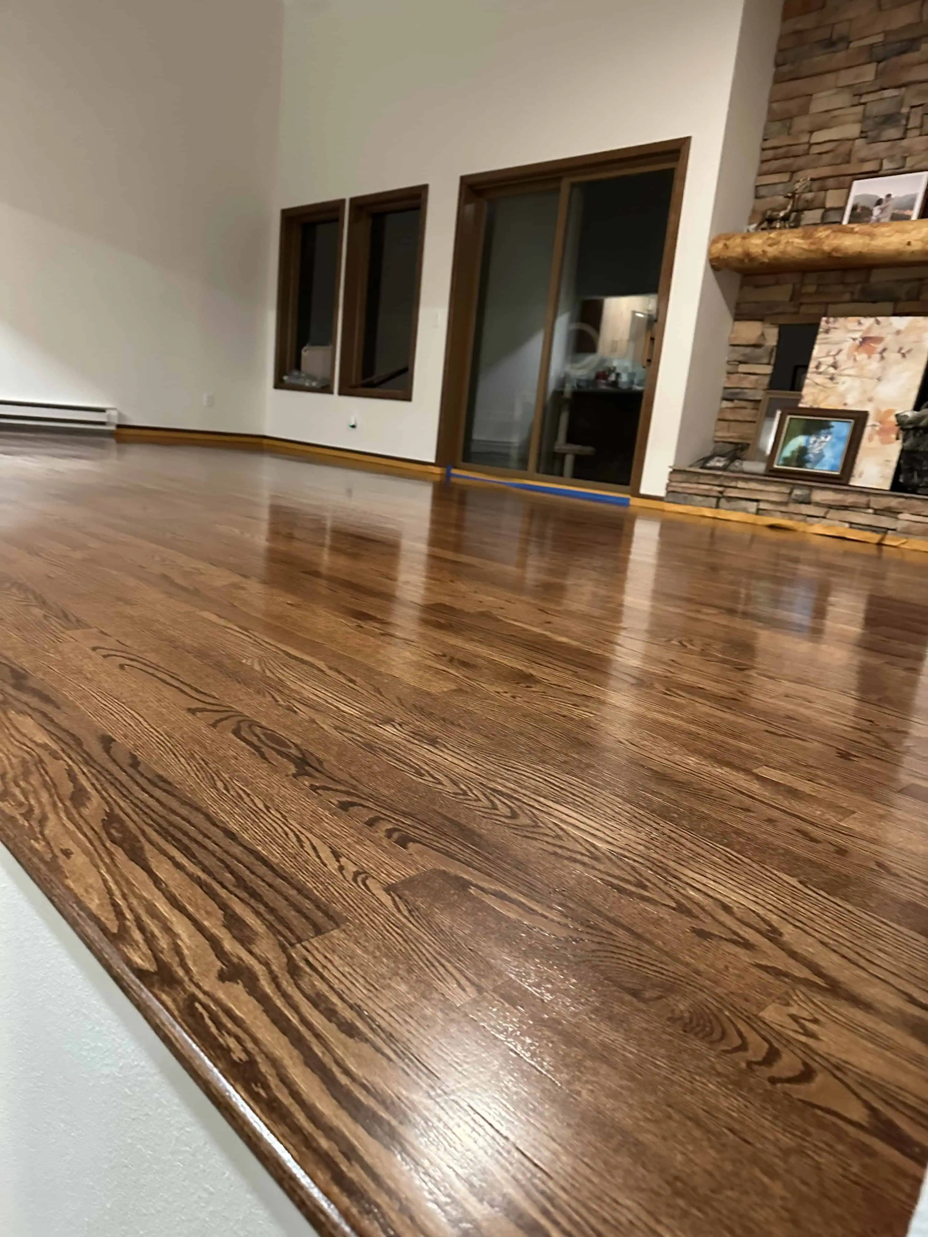 Newly refinished hardwood floor in a Denver home, featuring a rich, glossy finish with a rustic stone fireplace and large windows in the background, showcasing Blue Mountain Remodeling's craftsmanship in home renovation.