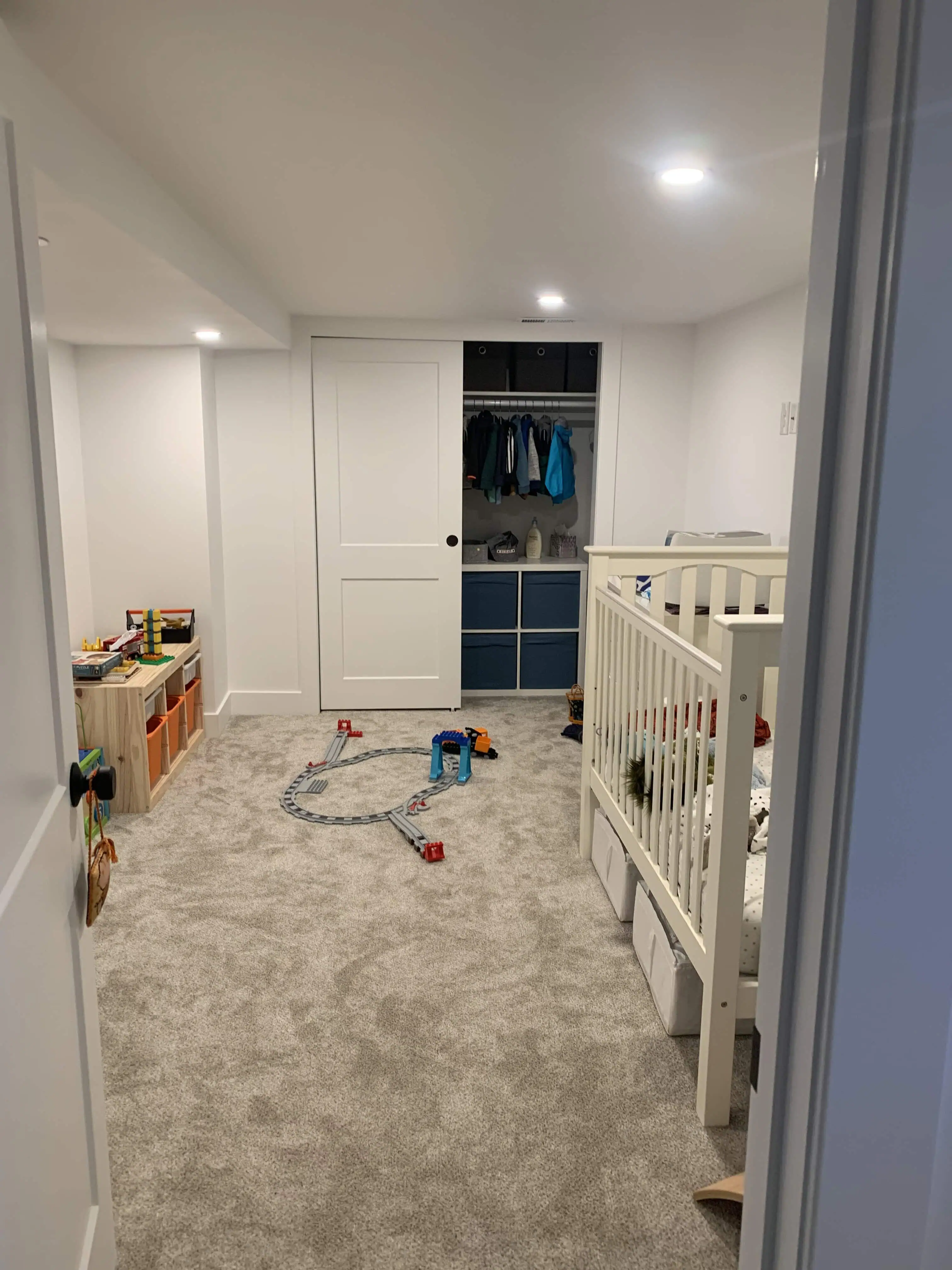 Renovated children's room with white walls, soft gray carpet, a crib, and a toy train set on the floor. Features an organized closet with sliding doors, showcasing Denver home renovation by Blue Mountain Remodeling.