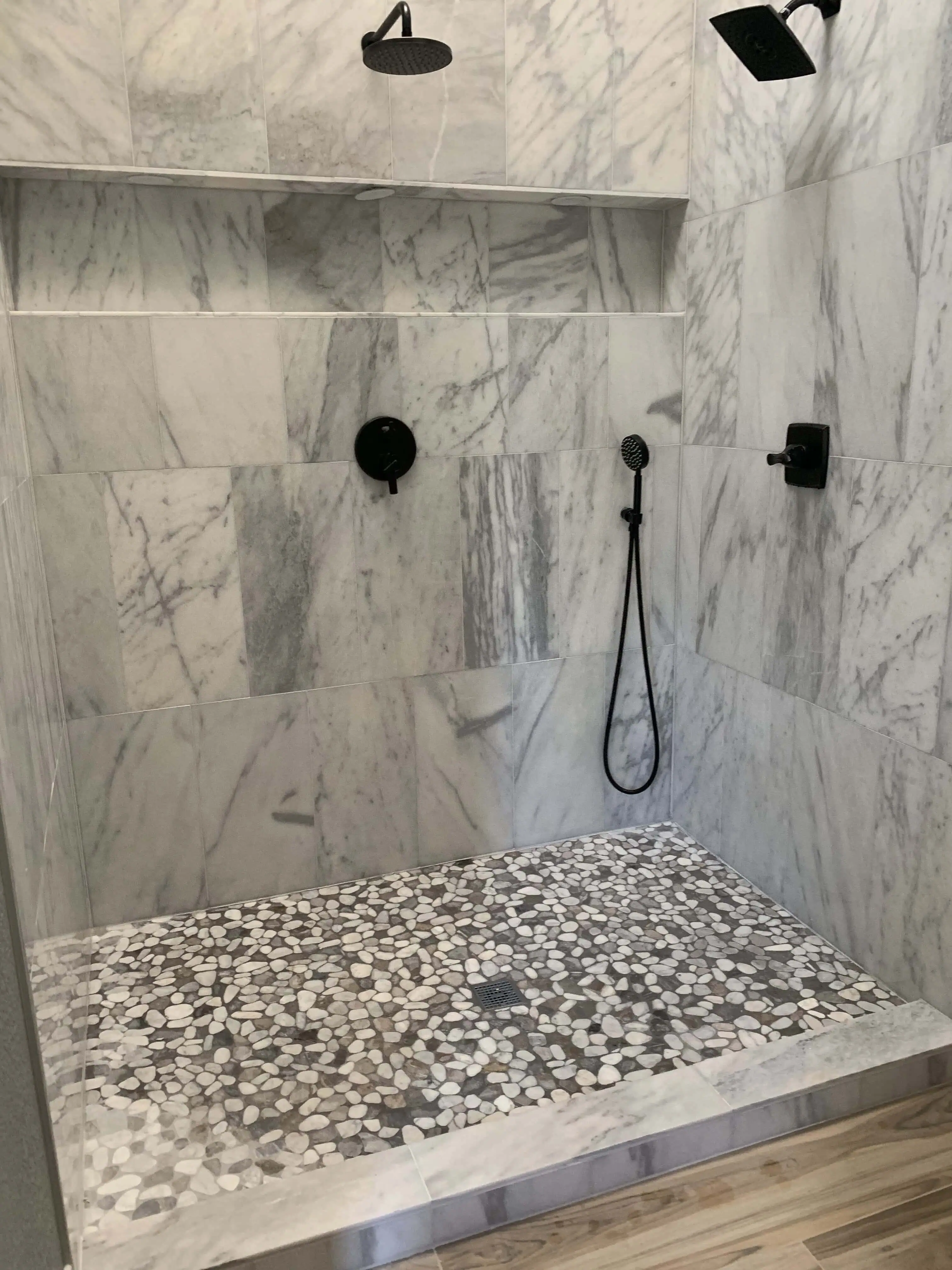 Luxurious marble shower with pebble stone flooring, featuring dual rainfall showerheads and a sleek handheld sprayer, by Blue Mountain Remodeling in Denver.