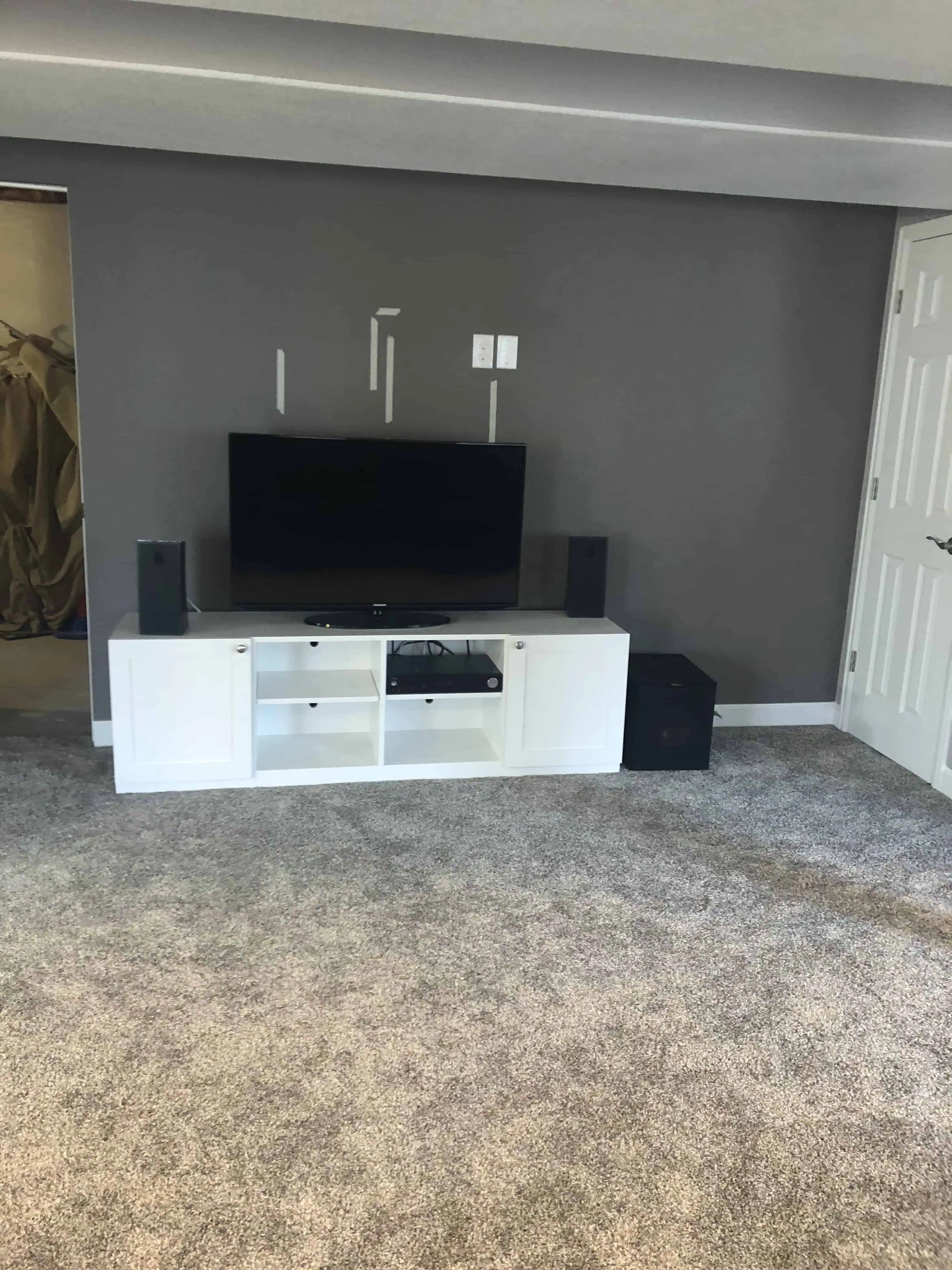 Modern living room with a flat-screen TV mounted on a gray wall and a white entertainment unit below. The room has gray carpeting and a closed door on the right. The space reflects Blue Mountain Remodeling's home renovation expertise in Denver.