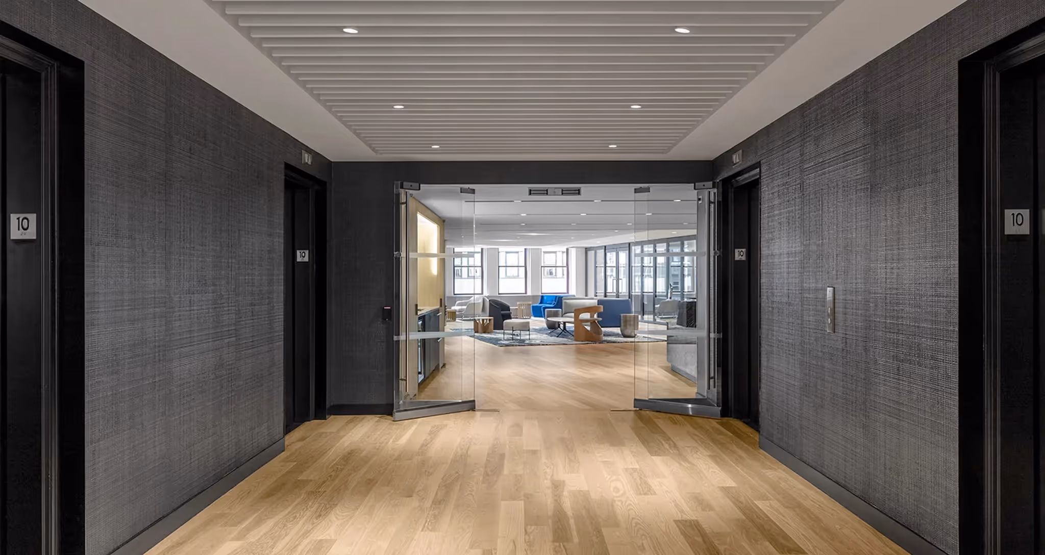 Office hallway with open glass doors leading to a modern lounge with wood floors and seating.