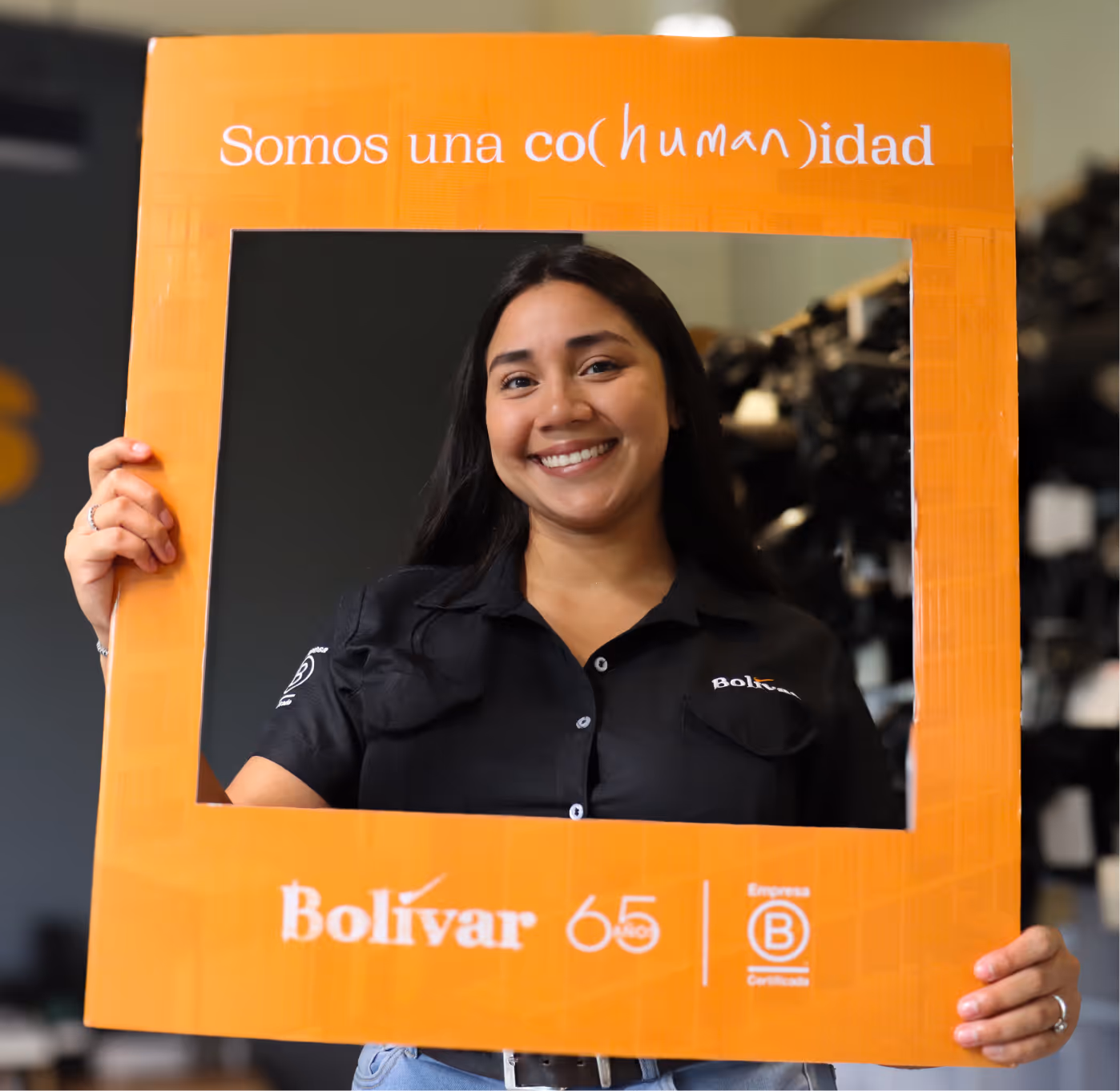Mujer sonriente sosteniendo un marco naranja con la frase 'Somos una co(human)idad' y logos de Bolívar 65 y Empresa B Certificada.