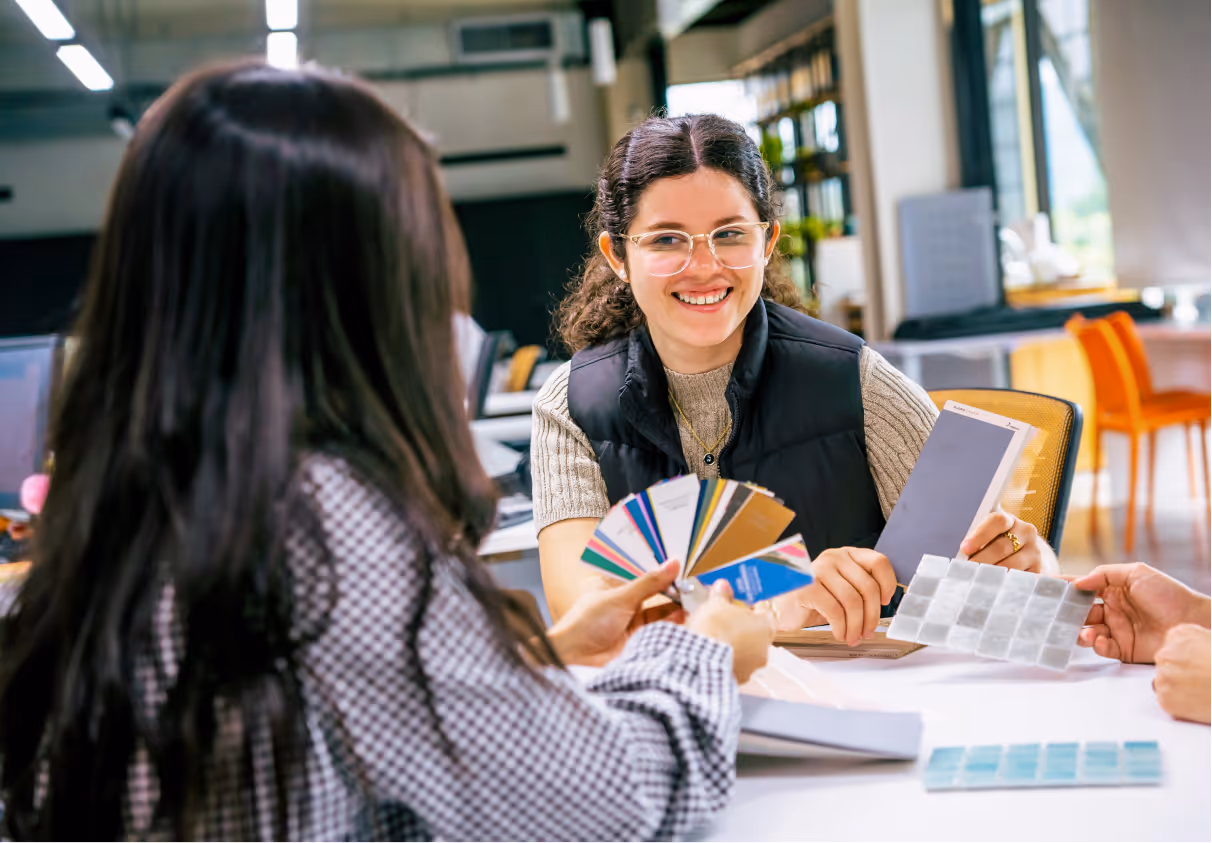 Dos mujeres sentadas en una mesa de trabajo revisan muestras de colores y texturas en una oficina moderna y luminosa.