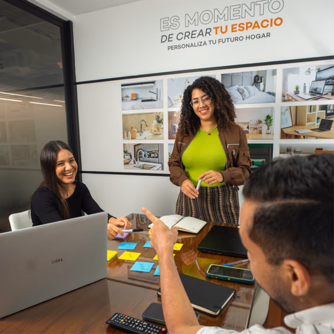 Tres personas sonrientes reunidas en una sala de reuniones con notas adhesivas y computadoras portátiles en la mesa.