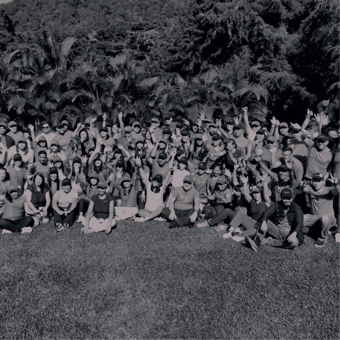 Gran grupo de personas, muchas con gorras, sonriendo y saludando mientras posan al aire libre con árboles y césped alrededor.