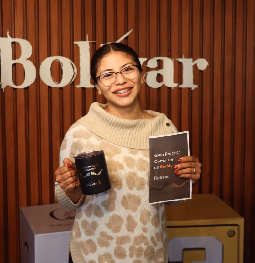 Mujer con gafas y suéter beige de estampado animal sonríe sosteniendo una taza negra y un folleto titulado 'Guía Práctica: Cómo ser un Buddy' frente a un fondo con la palabra Bolívar.