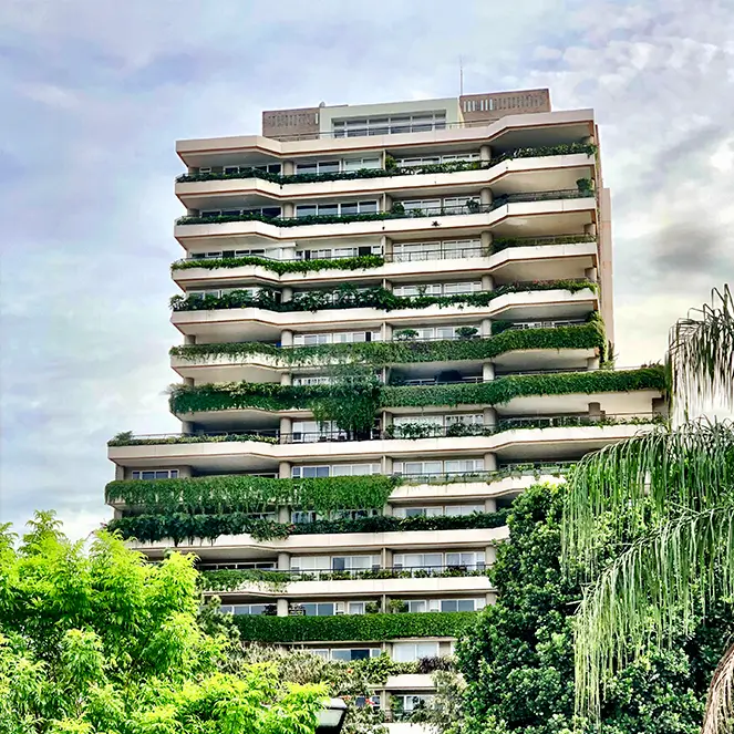 Edificio moderno de varios pisos con balcones y ventanas grandes frente a una colina cubierta de árboles y casas.