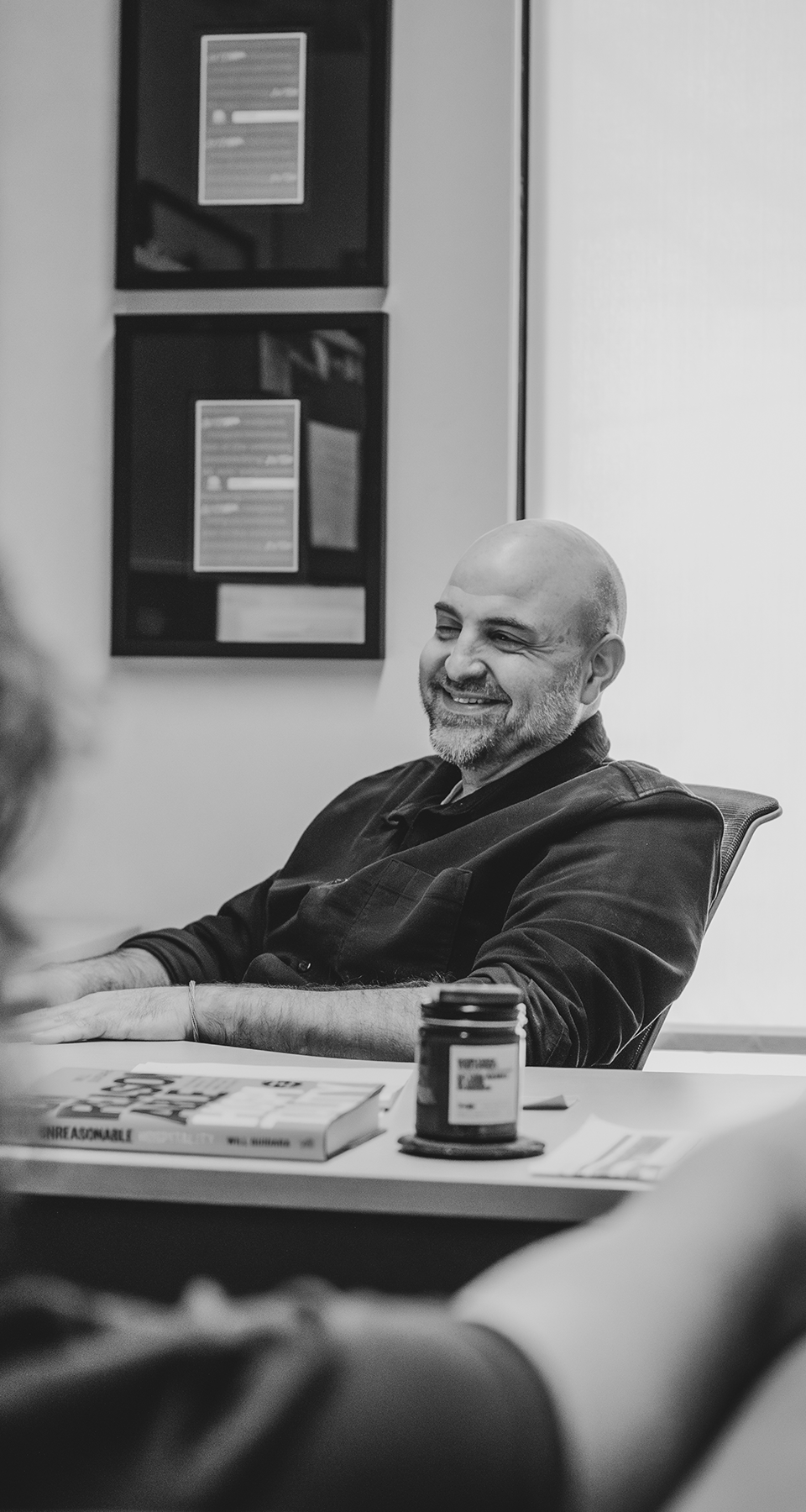 Man smiling and talking to a coworker while sitting at a desk in an office.