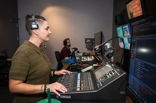 Technician adjusting controls on a Yamaha audio mixing console in a studio with multiple monitors and another person working in the background.