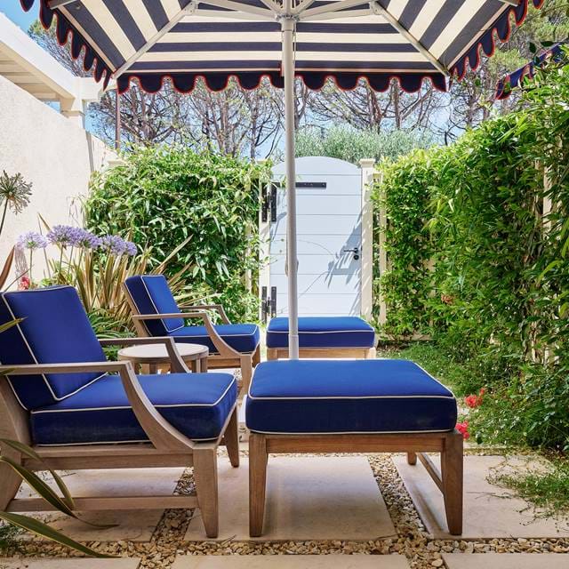 Private Terrasse im AREV Hotel in St. Tropez mit komfortablen marineblauen Liegestühlen unter einem gestreiften Sonnenschirm, umgeben von üppigem Grün.