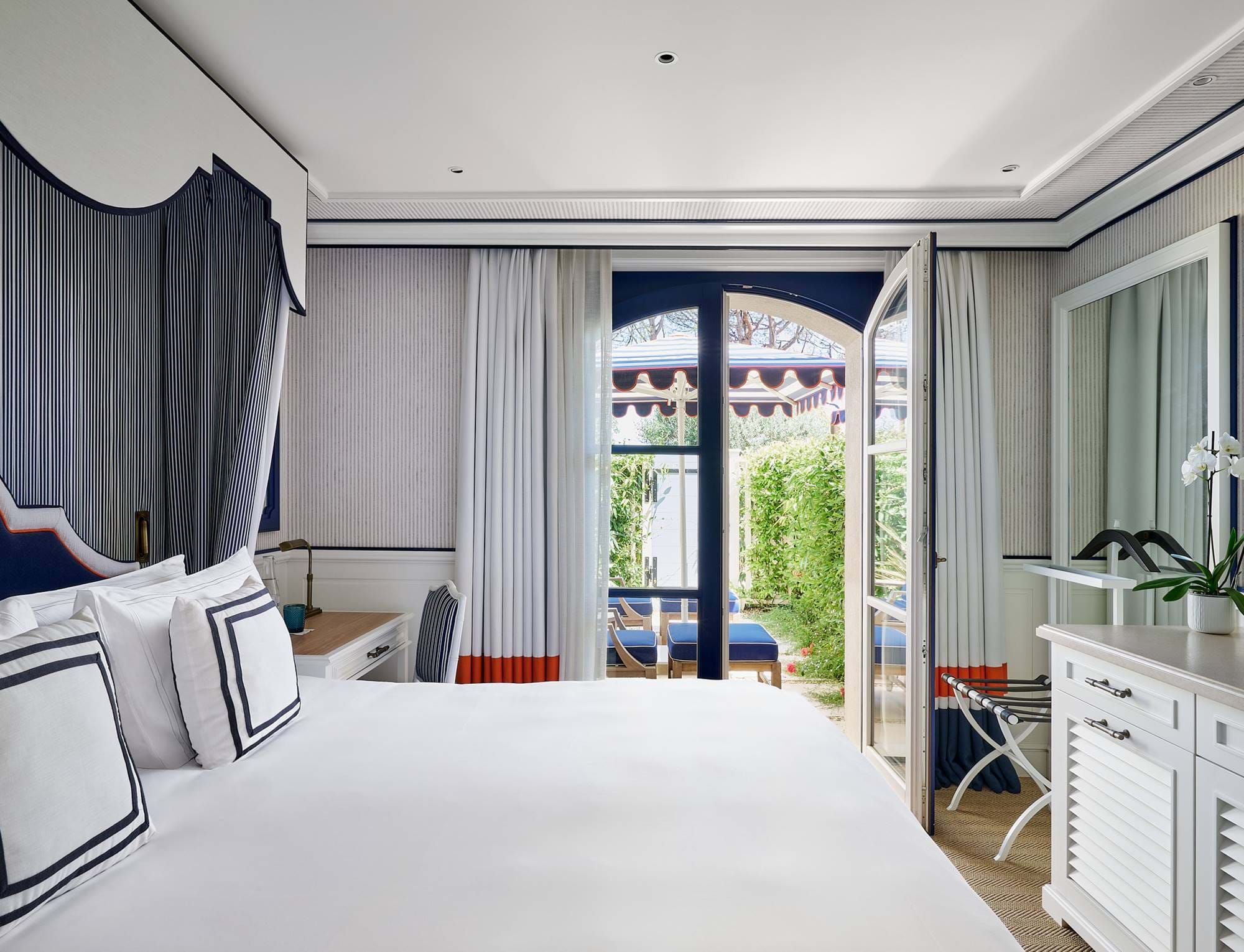 Helles Luxusschlafzimmer im AREV Hotel in Saint-Tropez mit Kingsize-Bett, elegantem weiß-blauem Dekor und einer offenen Tür, die zu einer privaten, blumengeschmückten Terrasse führt.