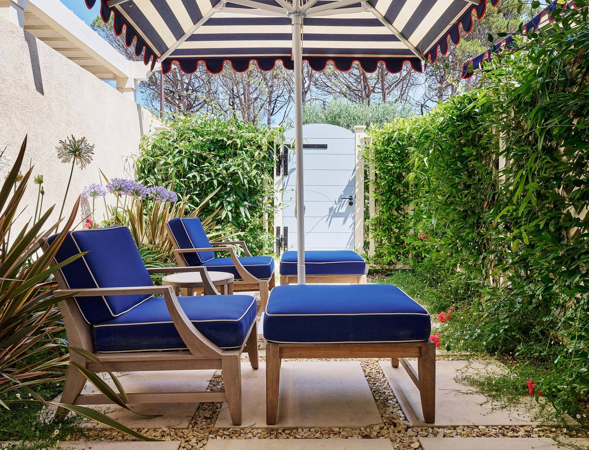 Private Terrasse im Hotel AREV in Saint-Tropez mit bequemen marineblauen Liegestühlen unter einem gestreiften Sonnenschirm, umgeben von üppigem Grün.