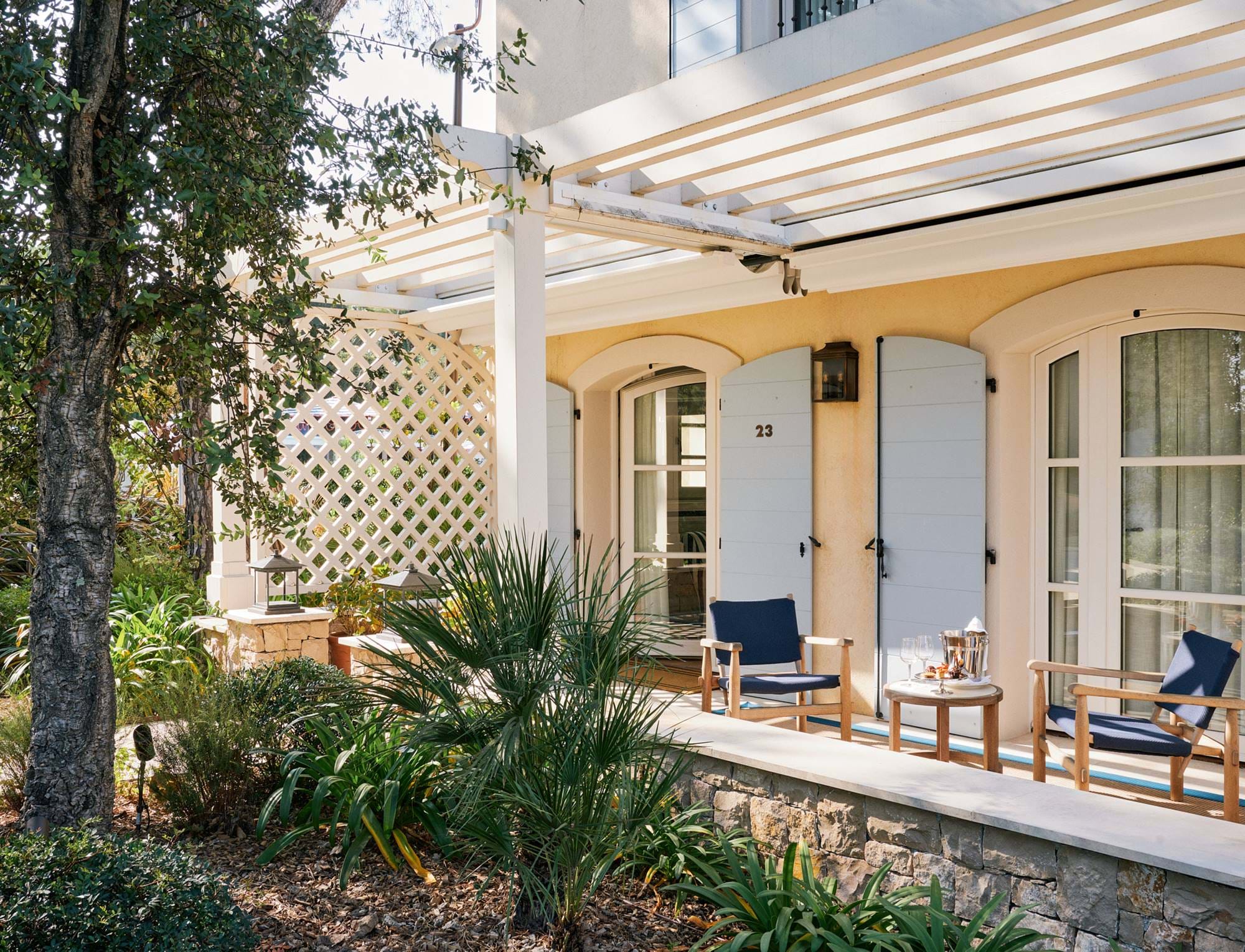 Sonnige Terrasse im Hotel AREV in Saint-Tropez mit bequemen Gartenmöbeln, einer weißen Pergola und üppigem Grün.
