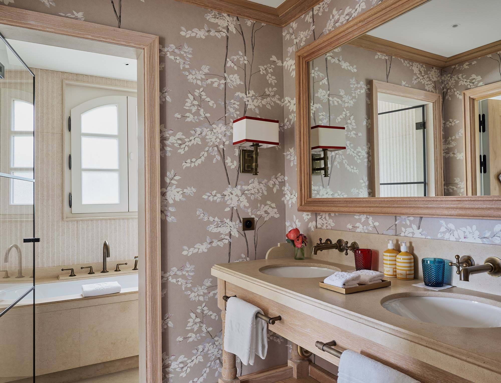 Banheiro sofisticado no hotel AREV em Saint-Tropez, com pia dupla de madeira, papel de parede estampado e banheira iluminada ao fundo.