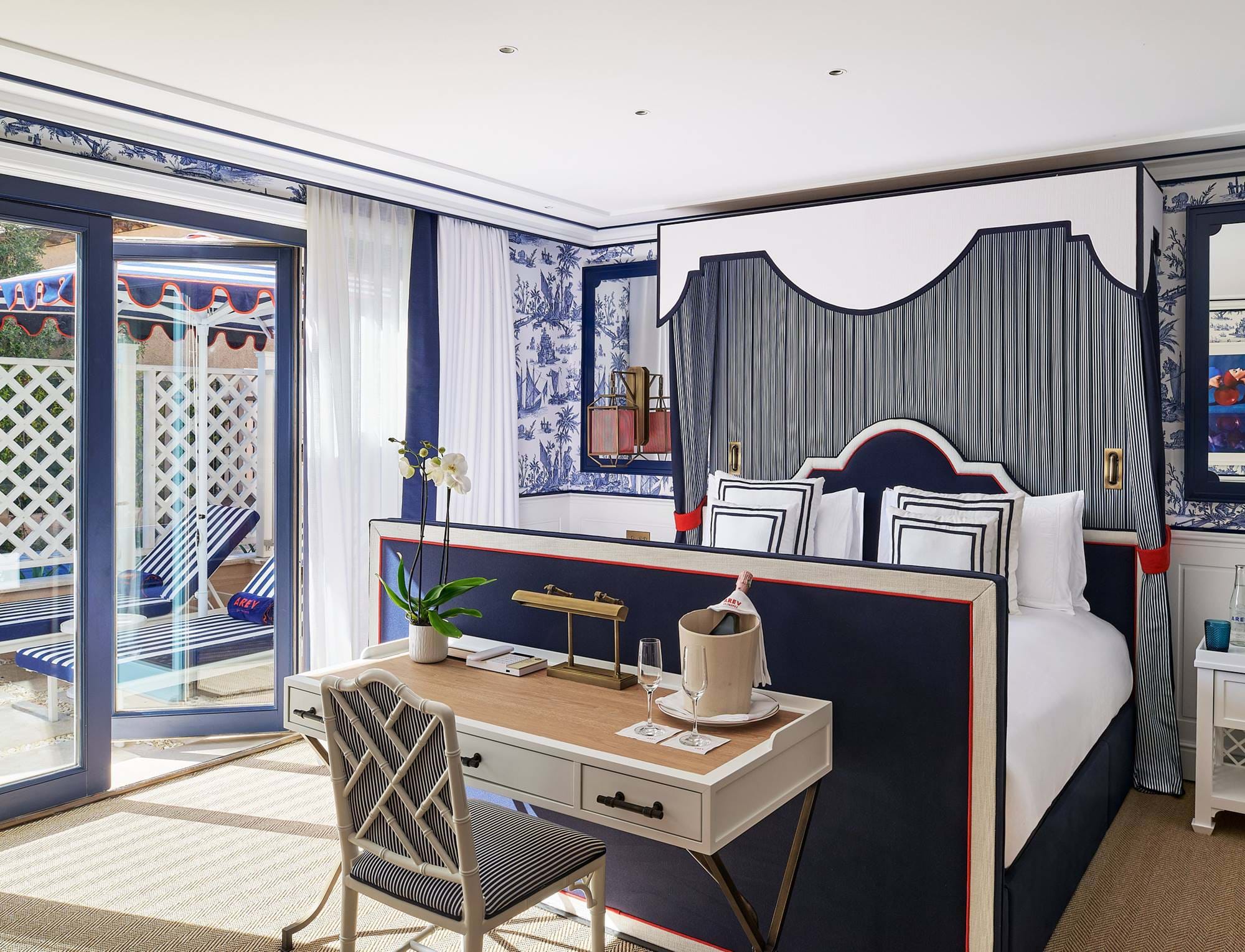 Innenansicht eines geräumigen Zimmers im AREV Hotel in Saint-Tropez mit Arbeitsbereich, marineblauer Bettwäsche und großen Glastüren.