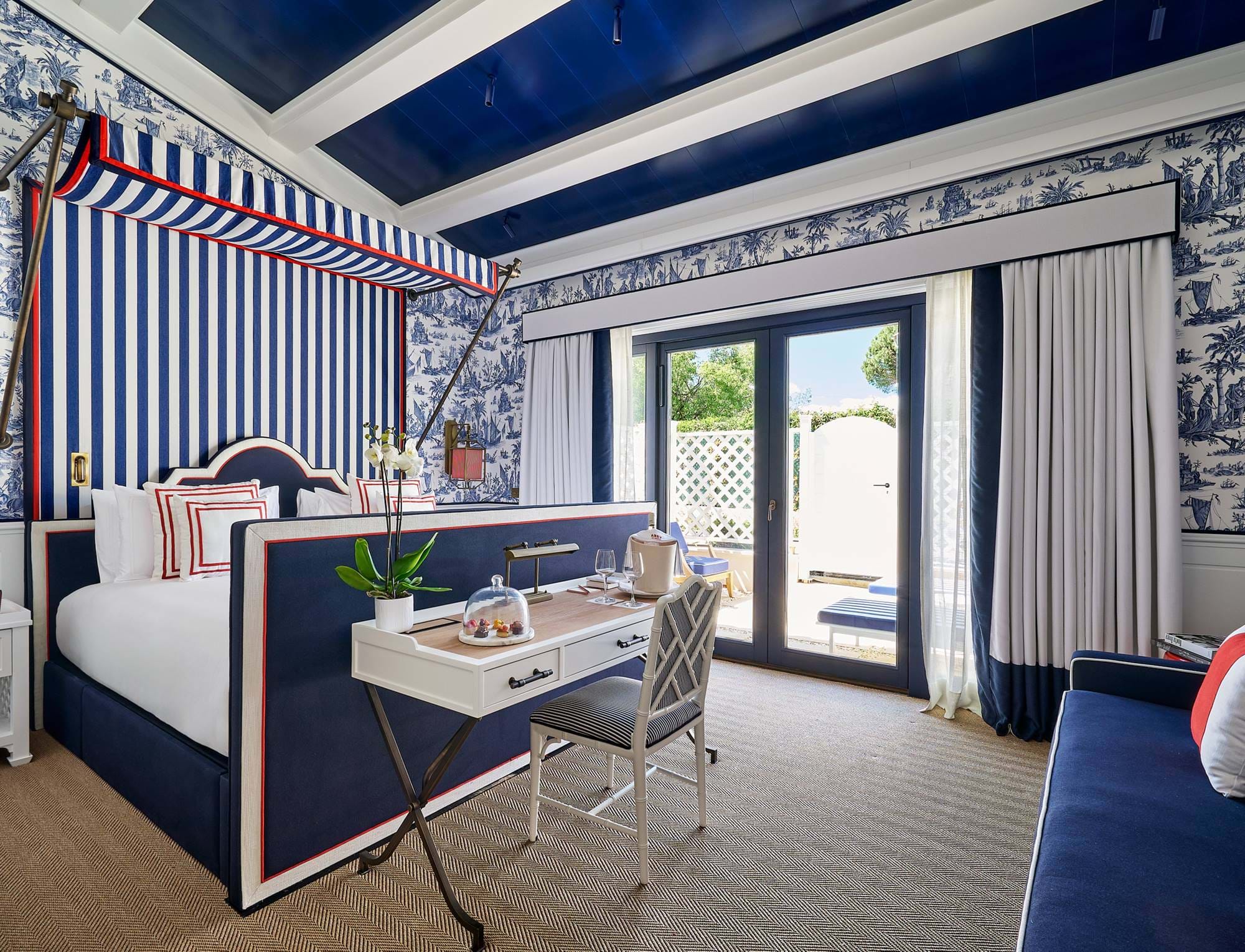 Luxussuite im AREV Hotel in Saint-Tropez mit einem blau-weiß gestreiften Himmelbett, einem eleganten Schreibtisch und Gartenblick.