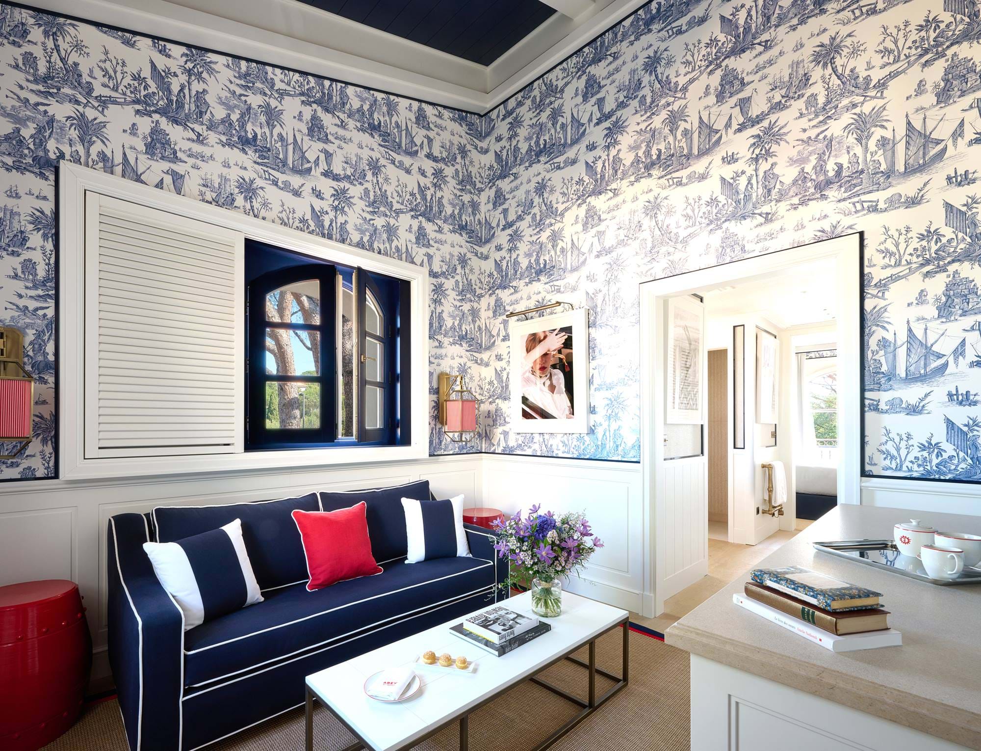 Schicker Wohnbereich im AREV Hotel in Saint-Tropez mit gemusterter Tapete, einem marineblauen Sofa und roten und weißen Dekorationselementen.